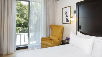 A hotel room with a bed, a yellow armchair, a small table with a vase of flowers, a framed picture on the wall, and a window with sheer curtains.