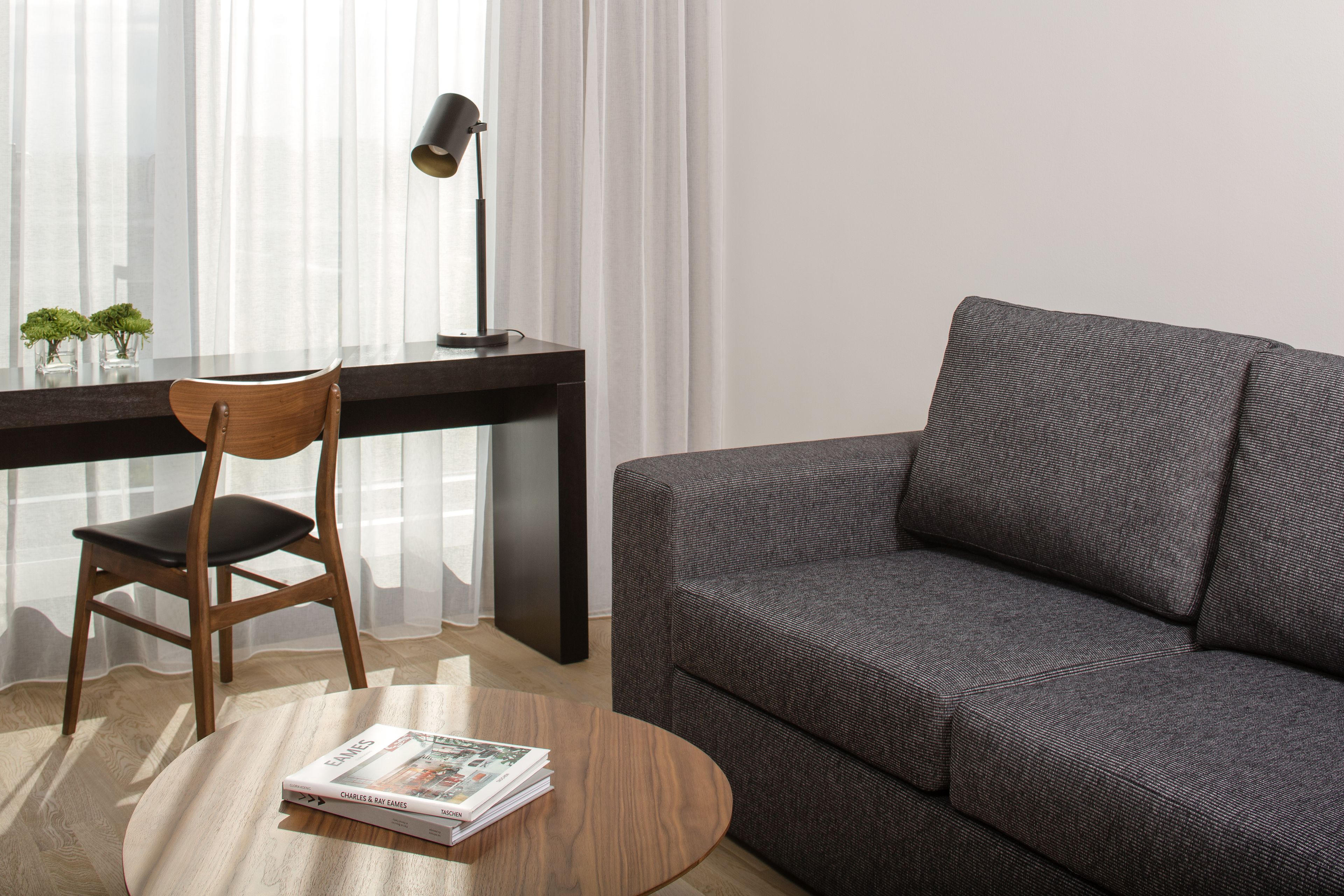 A modern living room with a dark grey sofa, a wooden chair, a round wooden coffee table with a magazine, and a desk with a lamp and two small potted plants.