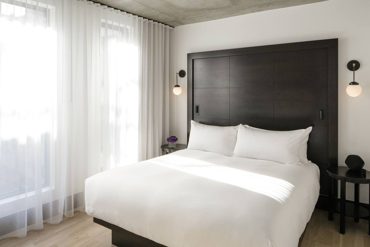 Une chambre d’hôtel moderne avec un grand lit, une tête de lit sombre, deux lampes de chevet et une fenêtre ornée de rideaux légers.