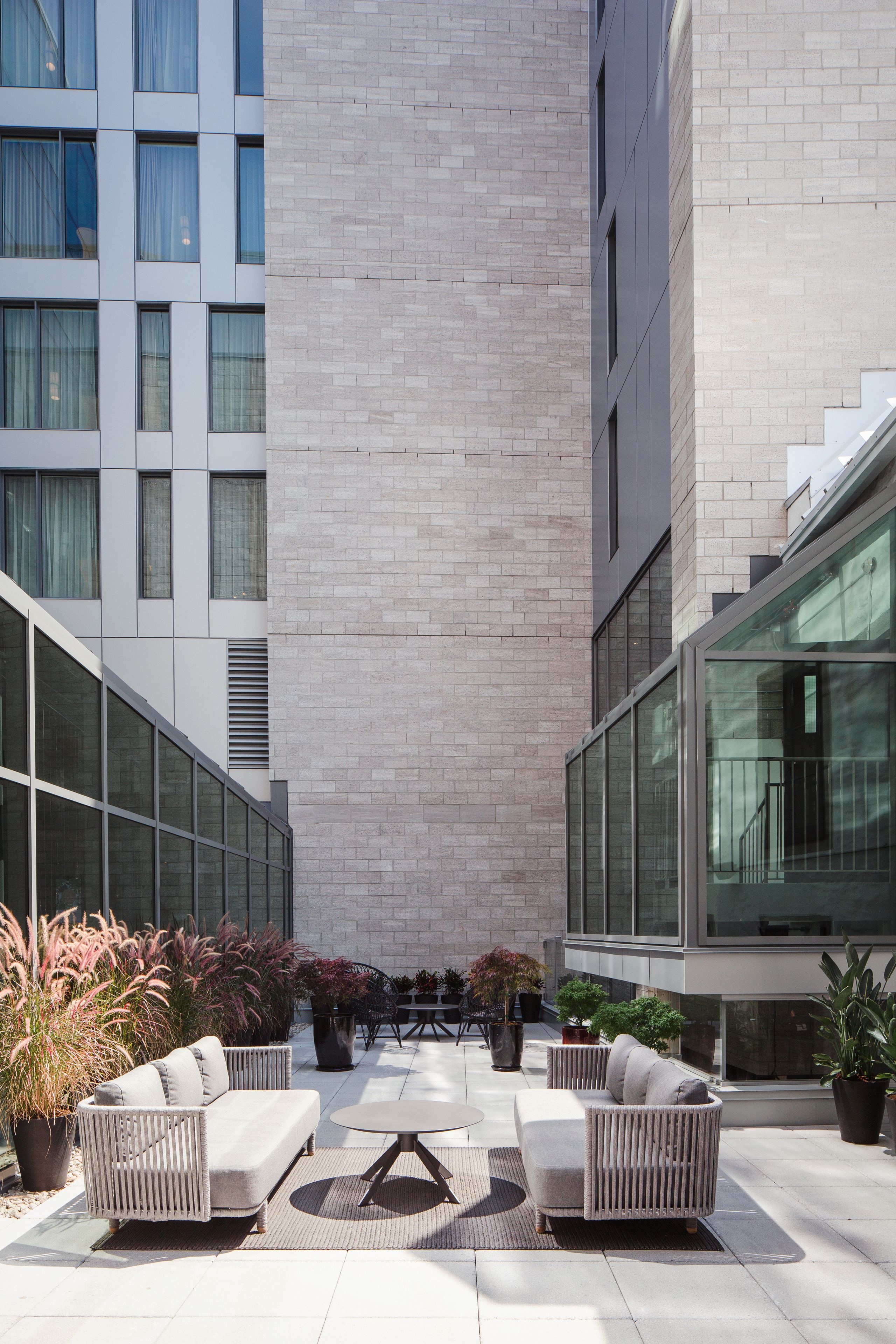 Un espace extérieur moderne aménagé avec deux canapés et une table, entouré de plantes en pot et d’un bâtiment doté de grandes fenêtres et d’un mur en briques.