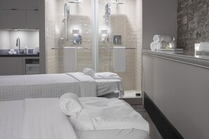 A modern hotel room with two beds, a glass-enclosed shower, and a bathroom with toiletries.
