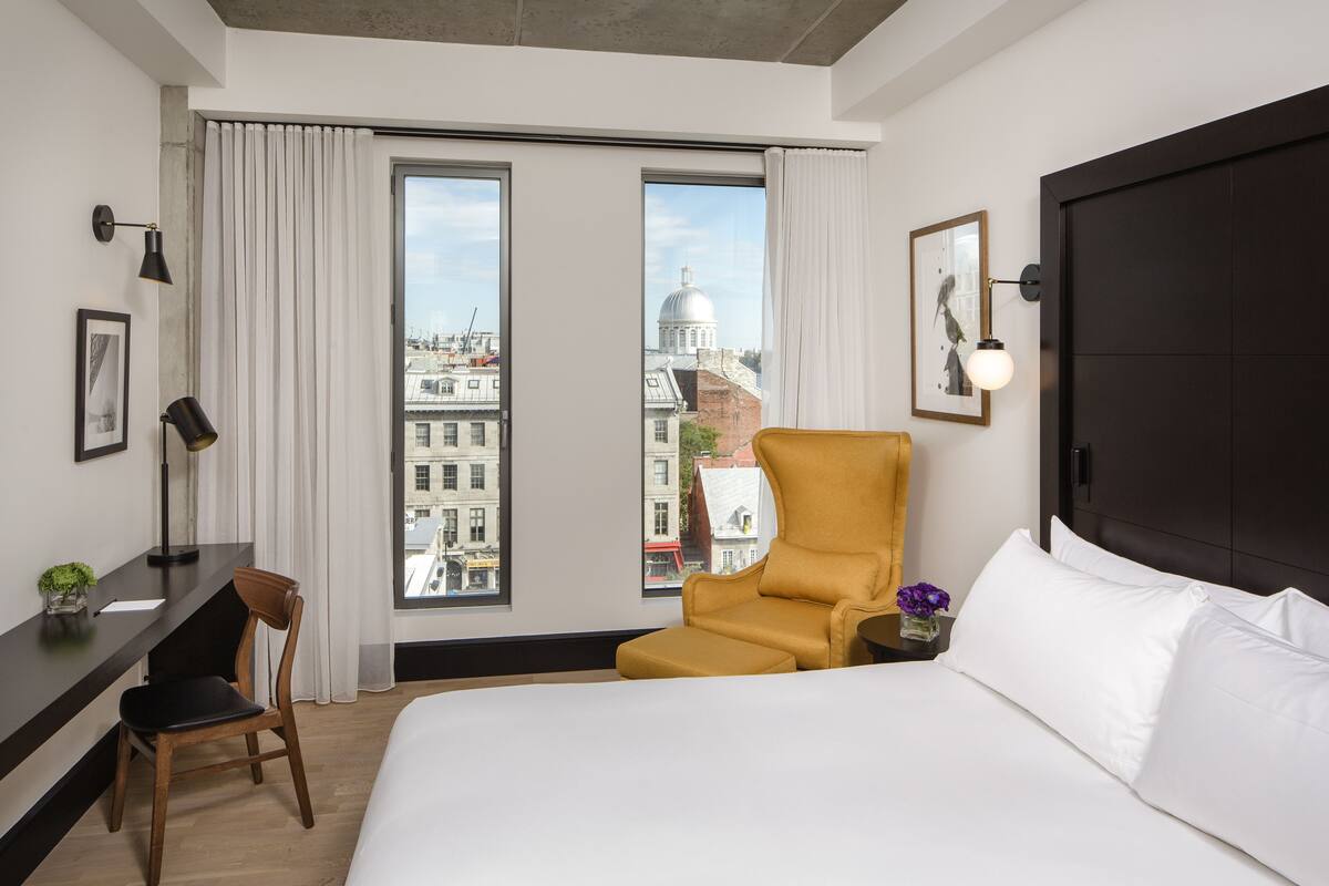 Une chambre d’hôtel avec un grand lit, un bureau avec une chaise, un fauteuil jaune et une vue sur les bâtiments de la ville à travers de grandes fenêtres.