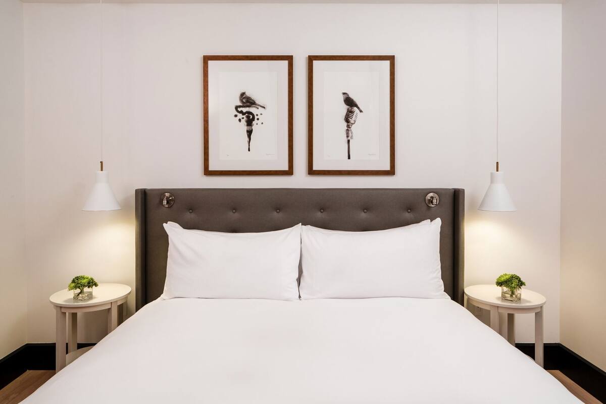 Un lit bien fait, doté d’une tête de lit capitonnée, avec deux œuvres d’art encadrées au-dessus, et deux tables de chevet ornées de lampes et de petites plantes en pot.