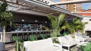 Ein Sitzbereich im Freien mit weißen Sofas und grünen Stühlen, eine Bar mit leuchtendem Neonzeichen und Blumentöpfen.