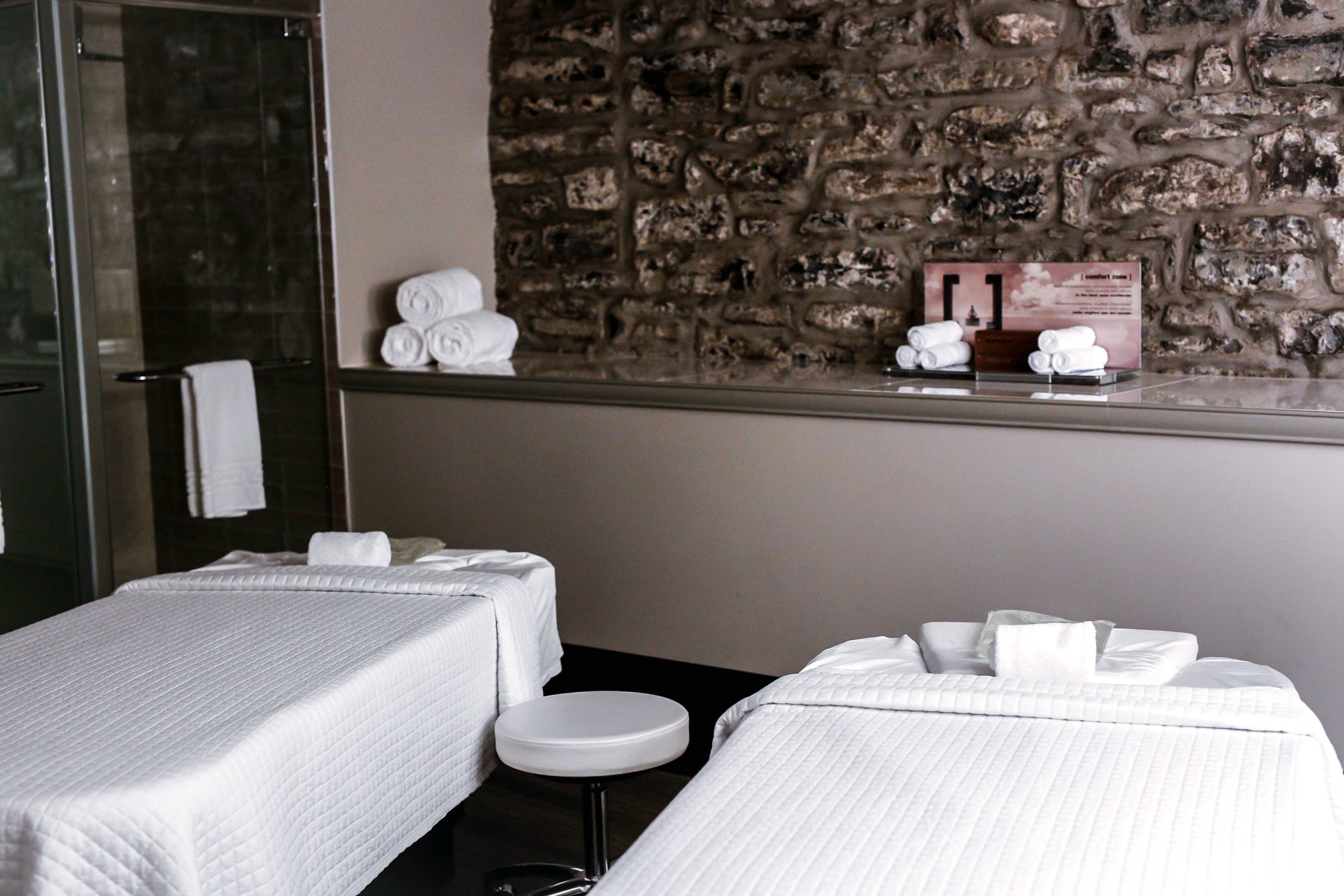 Habitación de hotel con dos camas, un taburete pequeño, pared de piedra y cabina de ducha de vidrio.