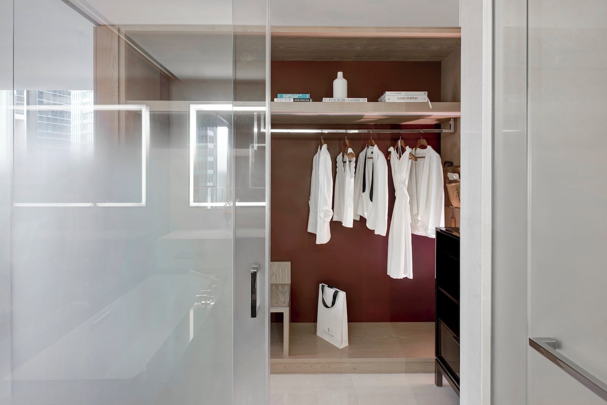 A modern closet with white robes hanging on a wooden rack, a white bag on the floor, and a small table with a chair.