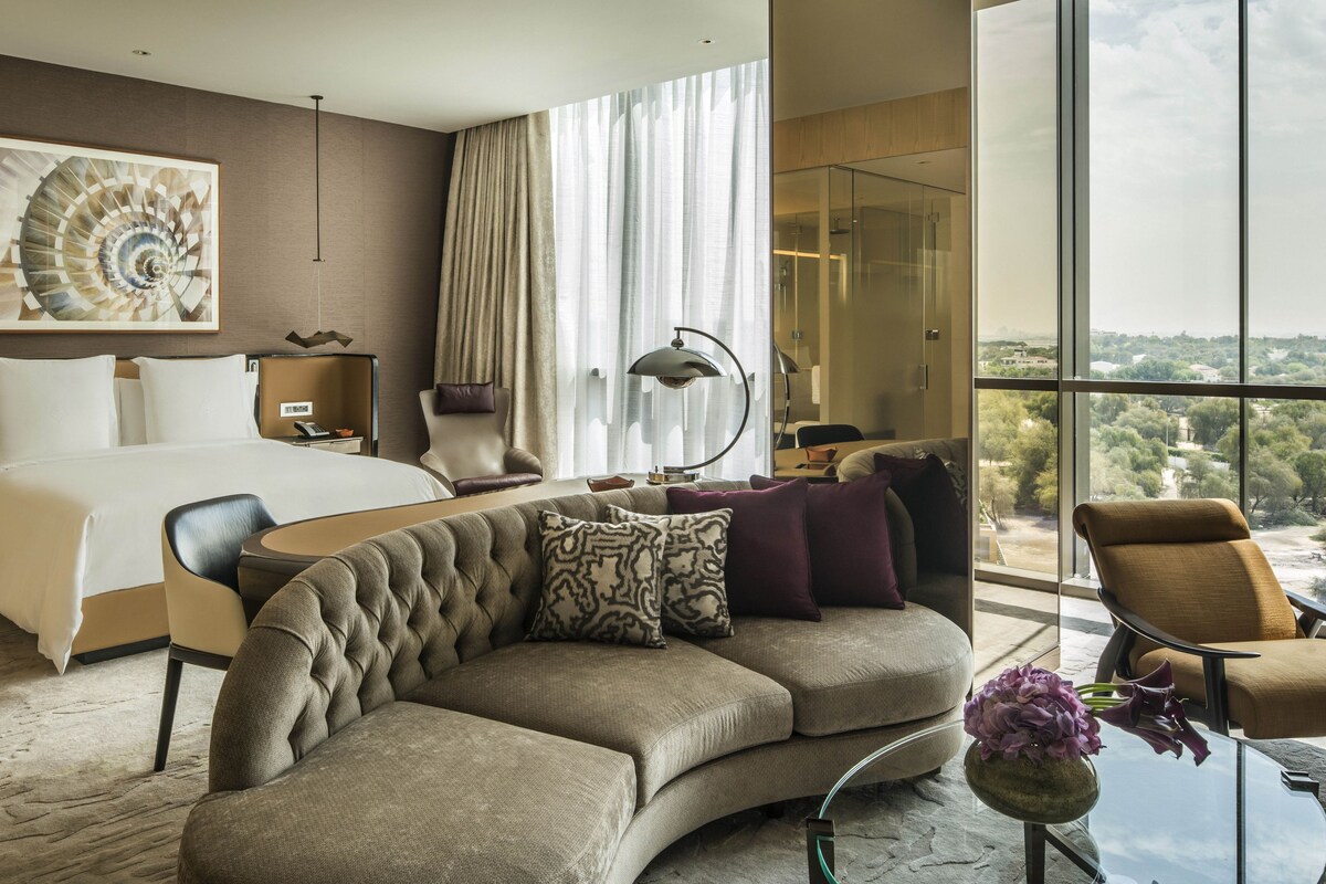 A modern hotel room with a large bed, a curved sofa, a desk, and a view of the city through large windows.