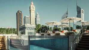 Un área de piscina en la azotea con sillones de descanso y sombrillas, rodeada de edificios modernos y una torre con reloj.
