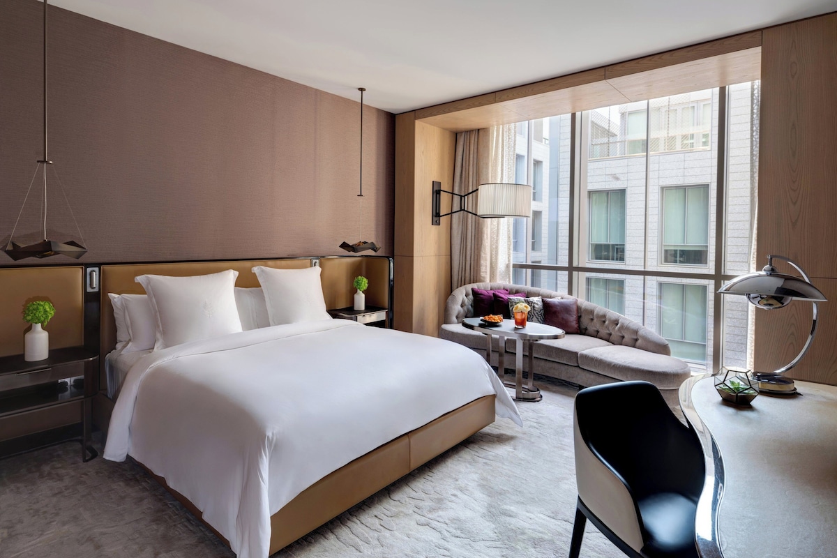 A modern hotel room with a large bed, a seating area with a view, and a small table with a drink.