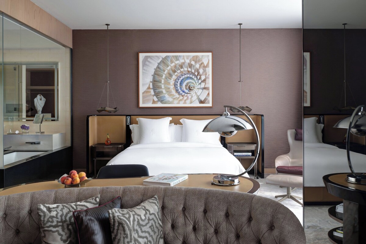 A modern hotel room with a large bed, a round desk, a sofa, a mirror, and a wall-mounted artwork.
