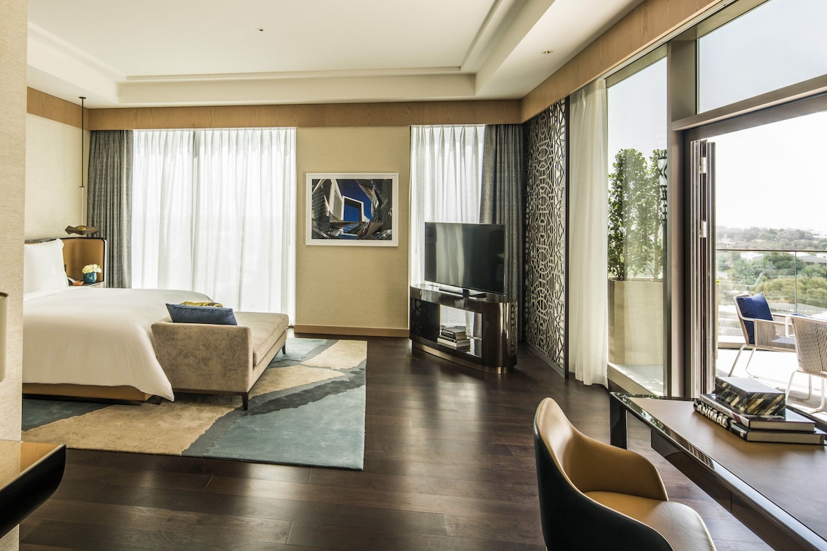 A modern hotel room with a large bed, a TV, a desk with a lamp, and a view of the city.