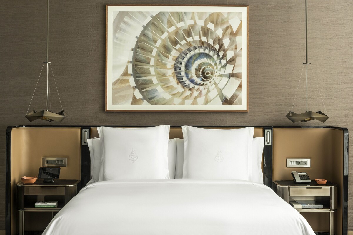 A hotel room with a bed, two nightstands, a wall-mounted artwork, and a hanging light fixture.