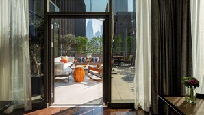 Puerta abierta que da a un balcón con mobiliario exterior, vistas de edificios altos y una planta en maceta sobre una mesita.