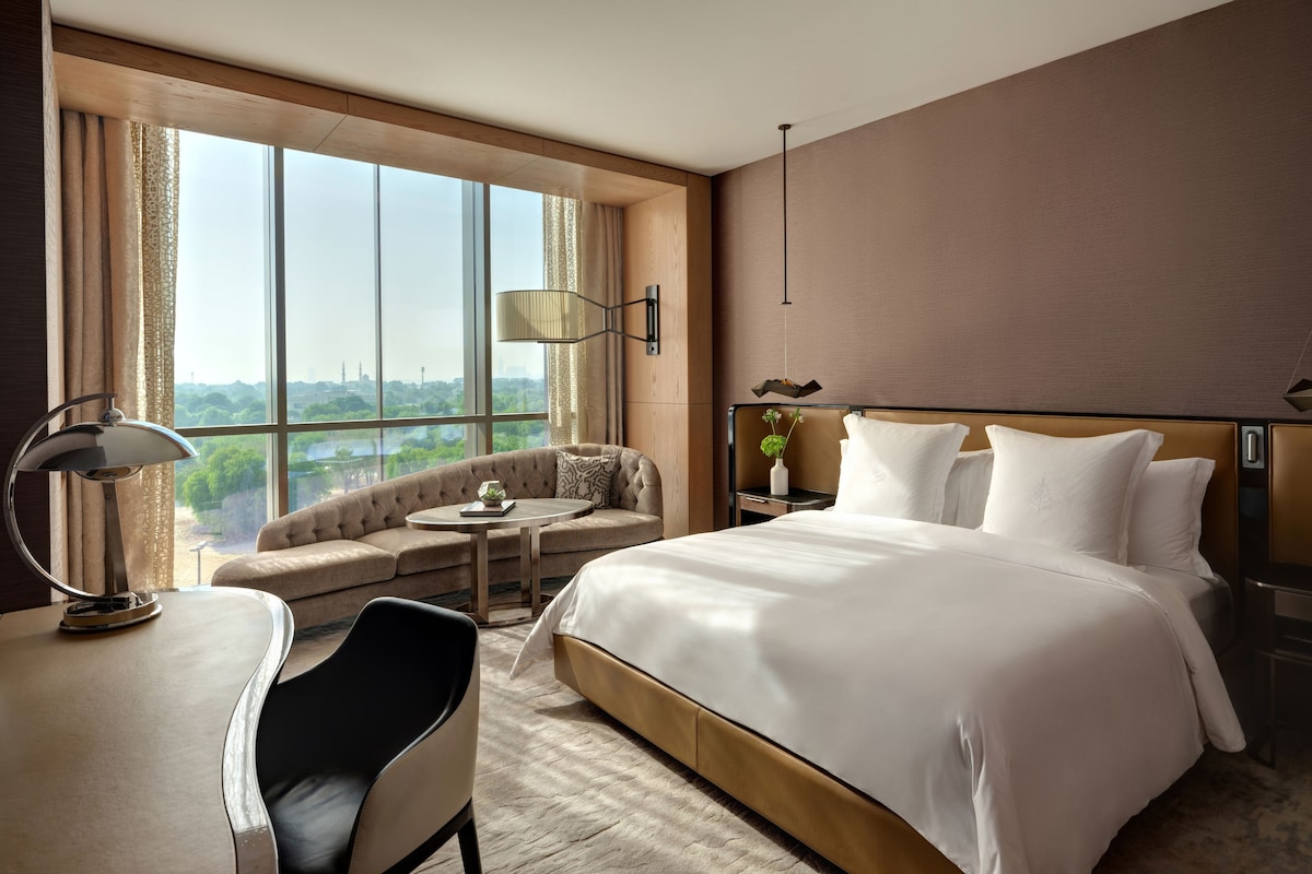 A modern hotel room with a large bed, a sofa, a desk, and a view of a cityscape through large windows.