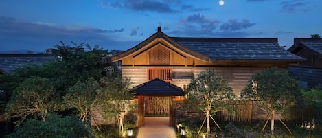 Una casa tradicional japonesa con fachada de madera, techo de tejas y un pequeño jardín con un sendero que conduce a la entrada.