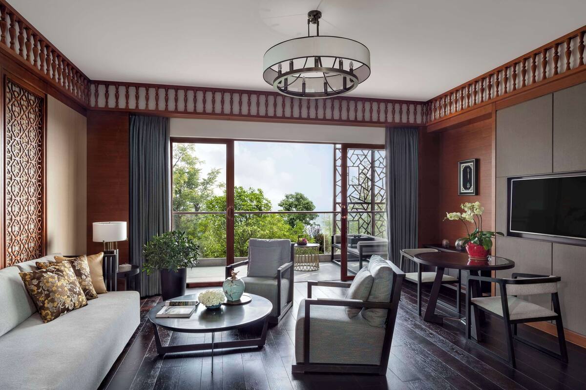 A modern living room with a sofa, coffee table, and chairs, a TV, and a balcony with a view of greenery.