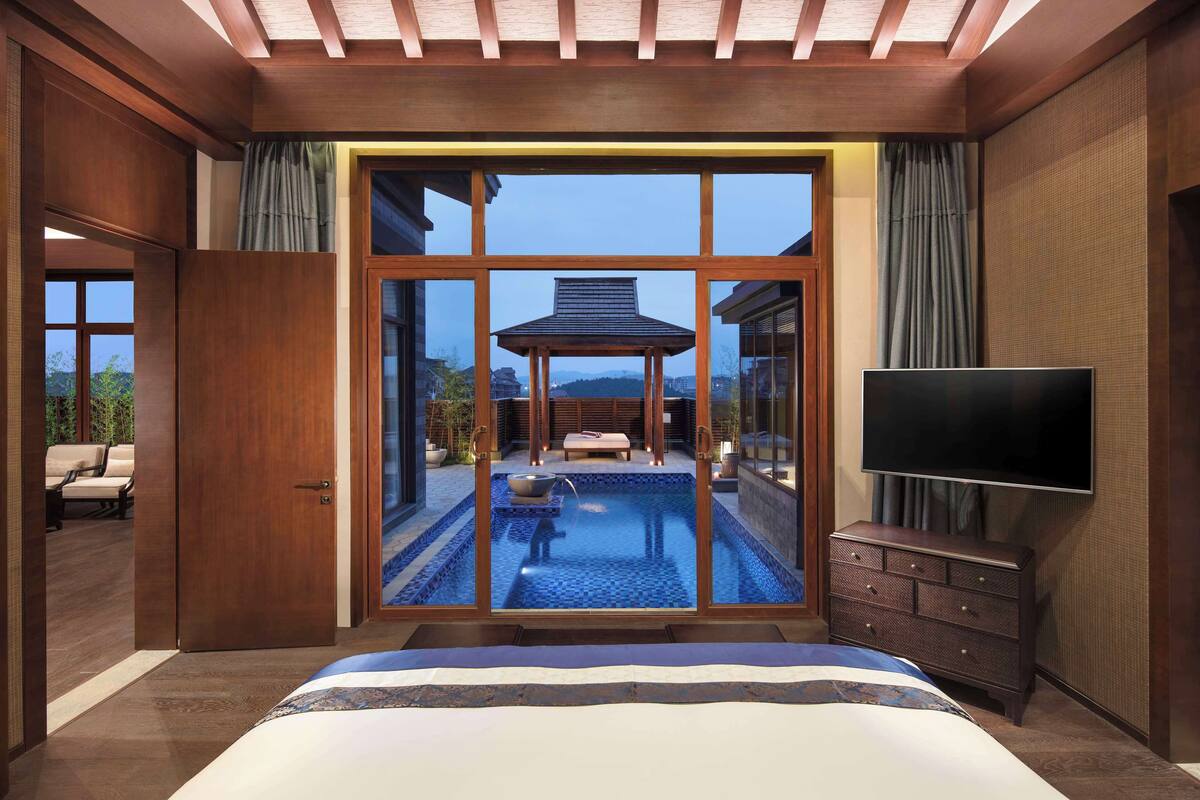 A bedroom with a large window, a TV, a wooden dresser, and a small pool.