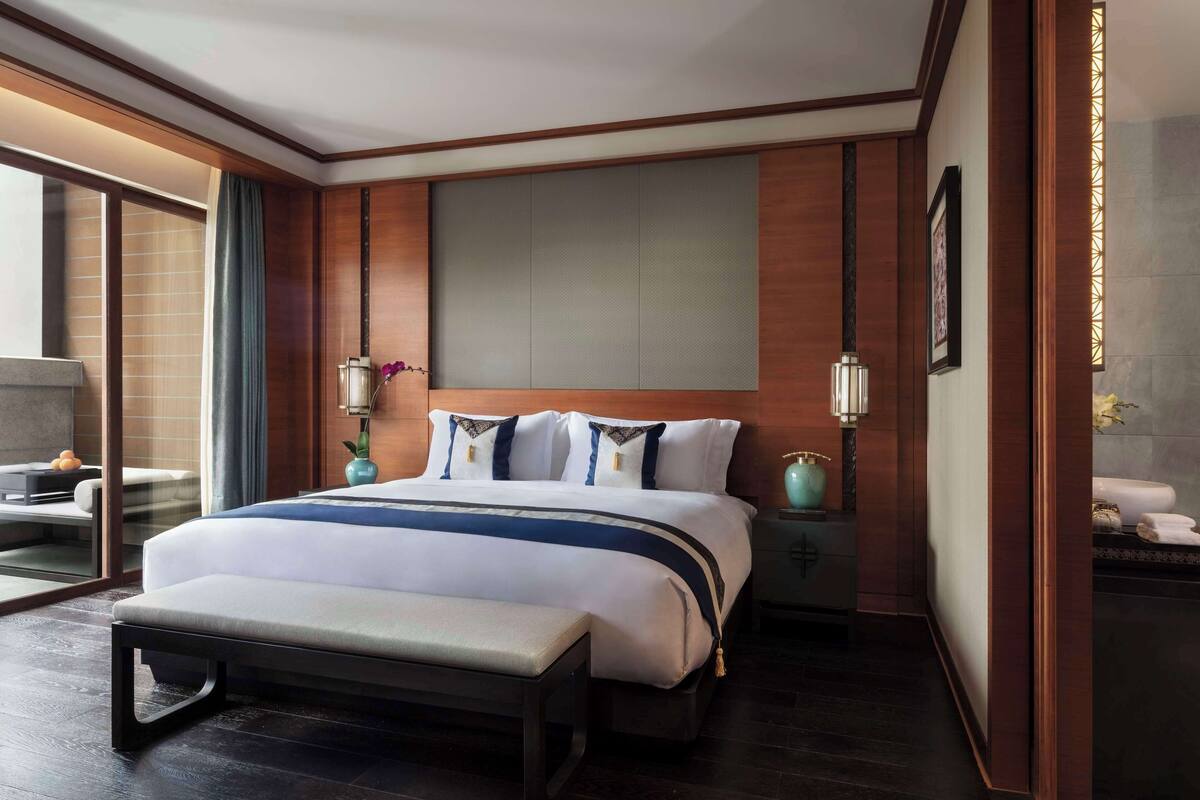 A modern hotel room with a large bed, a bench, a bedside table, and a large window.