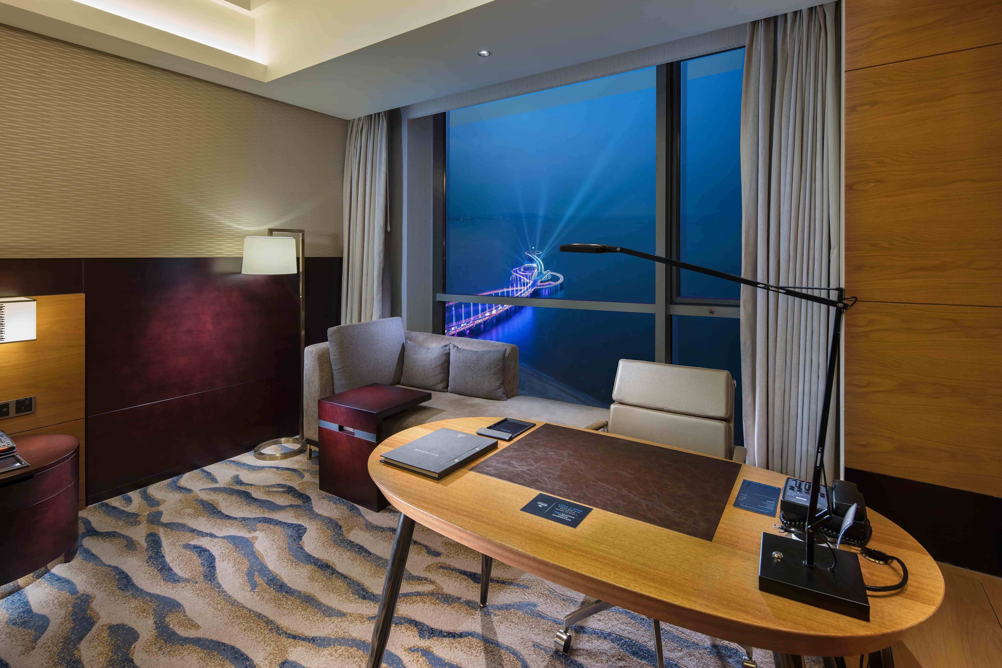 Une chambre d’hôtel avec une grande fenêtre, un bureau avec une lampe, un canapé et une petite table sur laquelle se trouve un ordinateur portable.