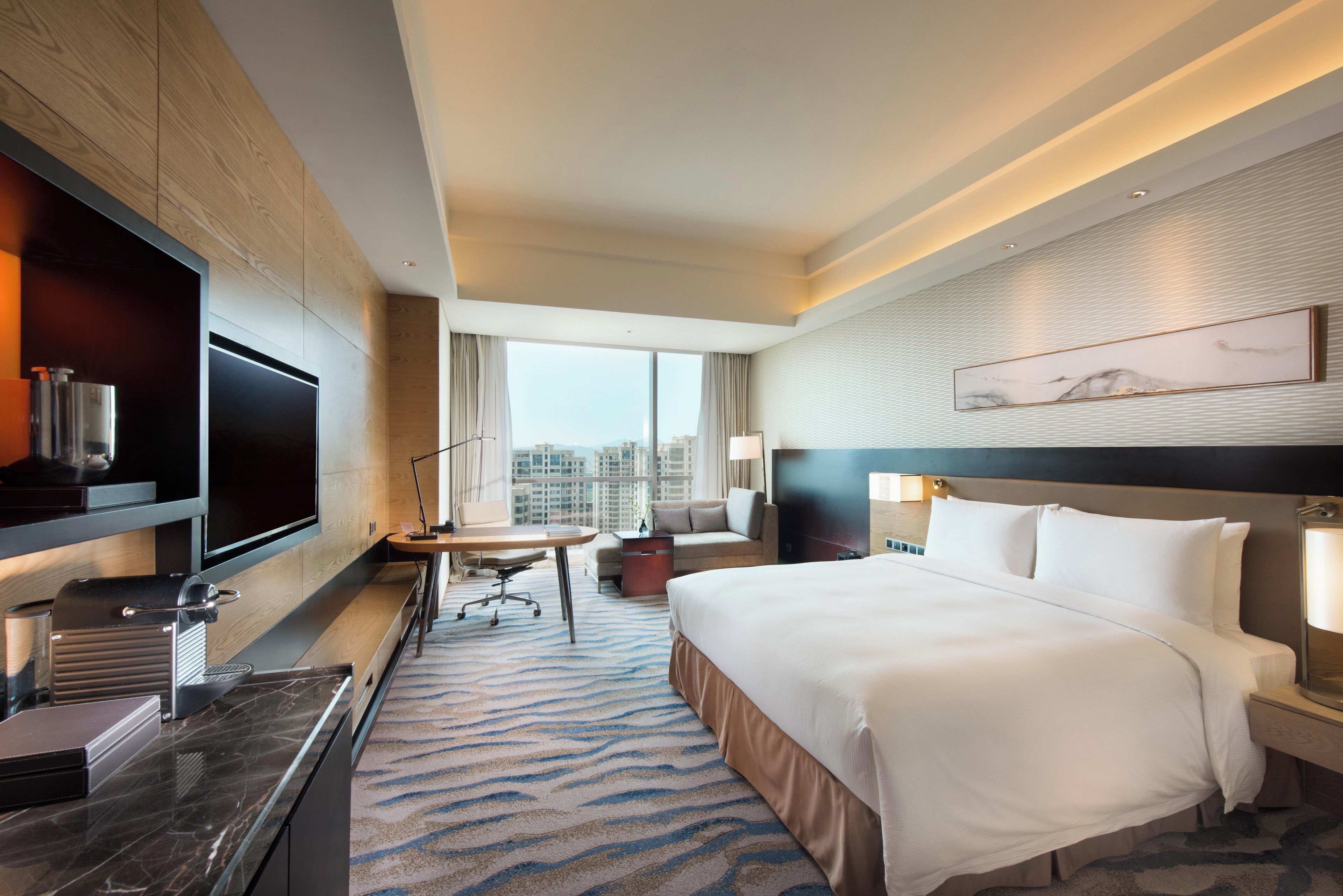 A hotel room with a large bed, a desk with a chair, a flat-screen TV, a minibar, and a city view.