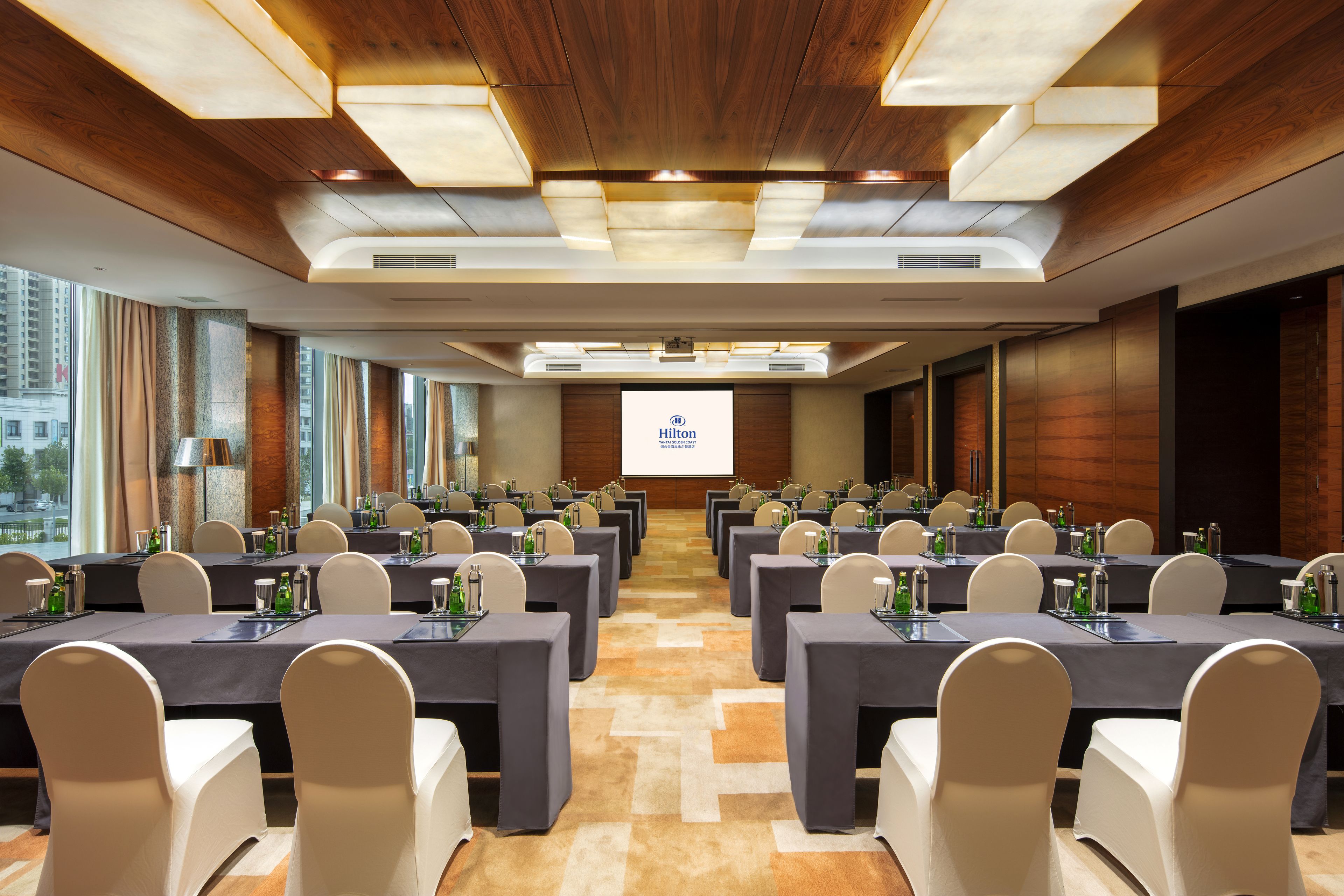 A conference room with multiple tables and chairs, a large screen at the front, and a view of the city through large windows.