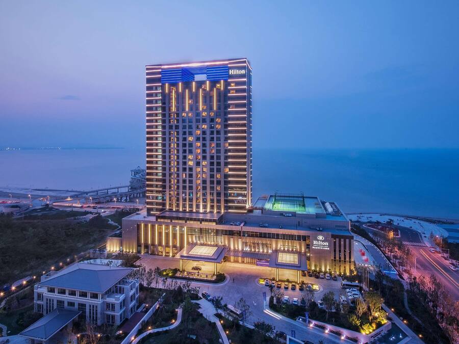 A modern hotel with a prominent Hilton sign, featuring a tall structure with illuminated windows and a well-lit entrance area.