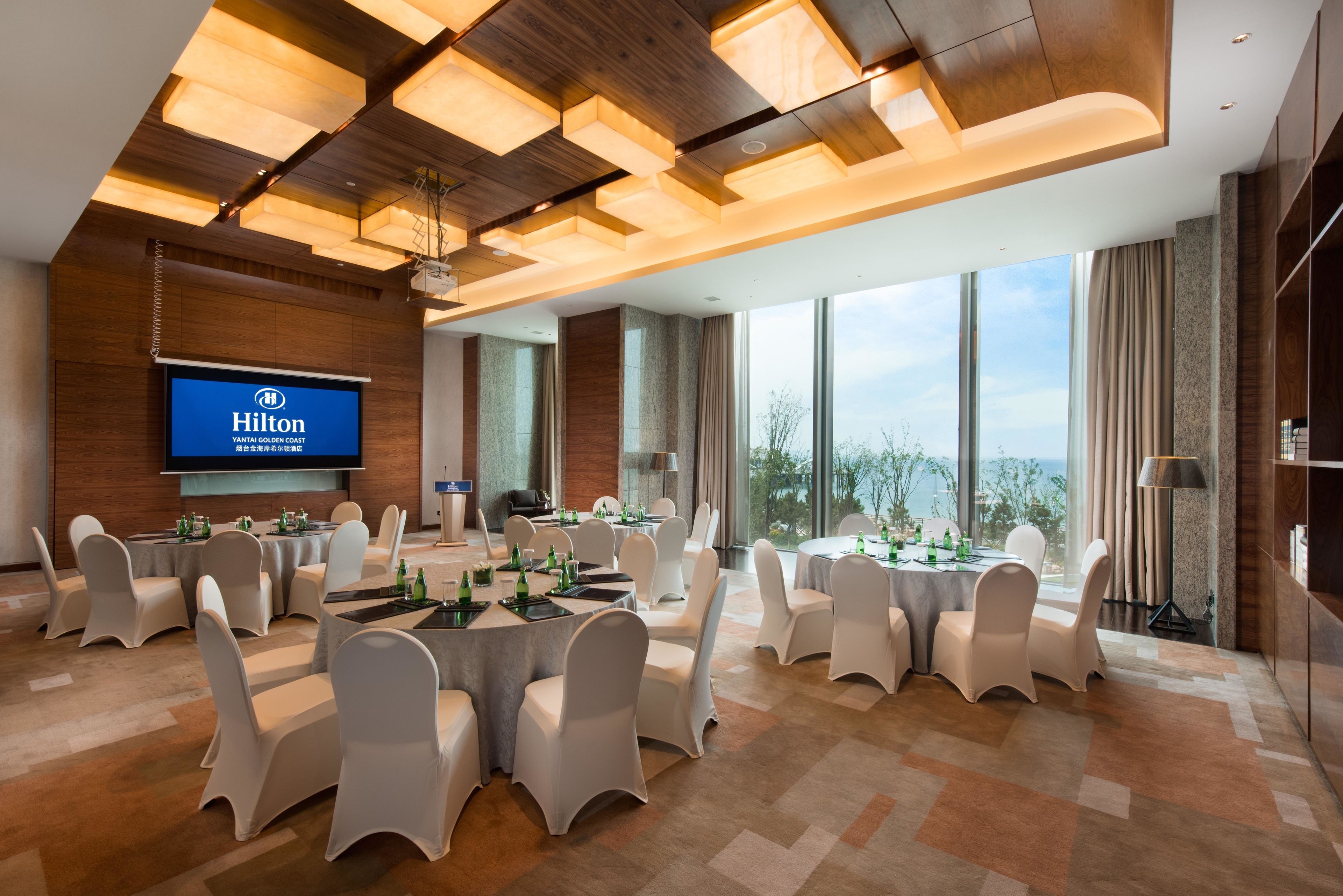 A conference room with round tables, white chairs, and a large screen displaying the Hilton logo.
