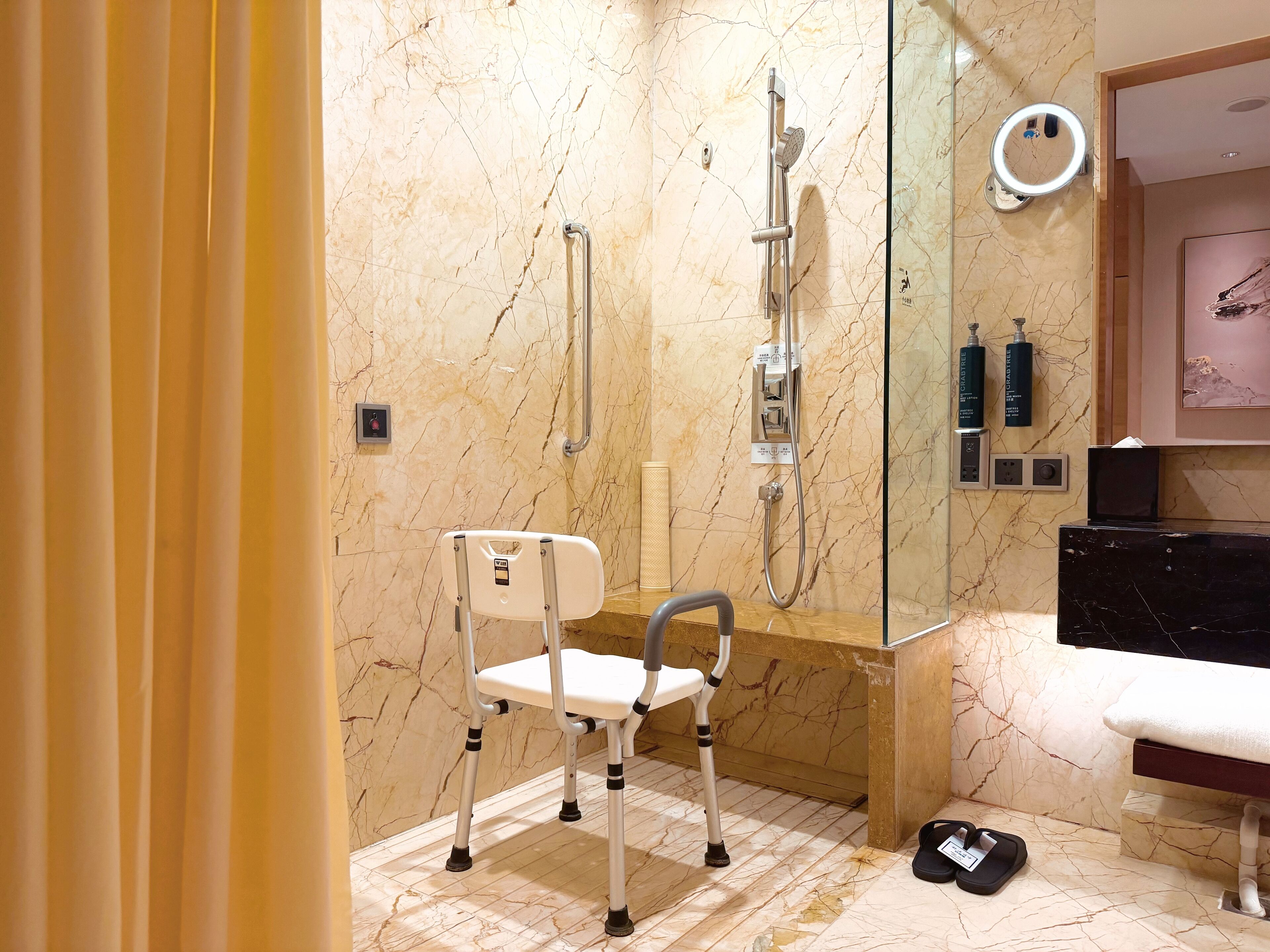 Un baño moderno con suelo de mármol, ducha con cabezal de mano, una silla blanca y un banco negro.