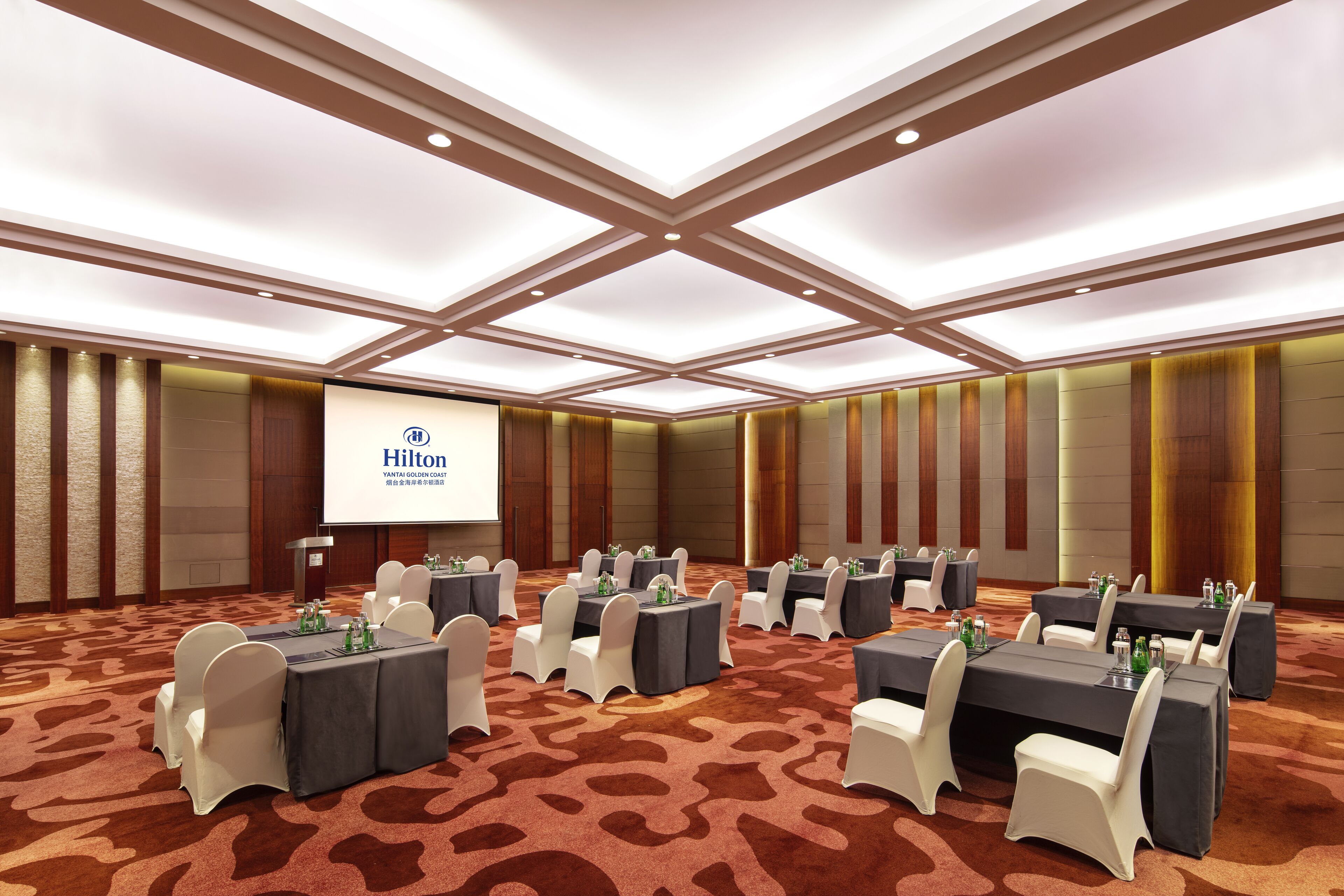 A conference room with multiple tables and chairs, a large screen displaying the Hilton logo, and a carpeted floor.