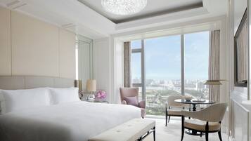 Amplia habitación de hotel con una cama grande, un espacio de descanso con una mesa y sillas, un banco y vistas de la ciudad a través de grandes ventanales.