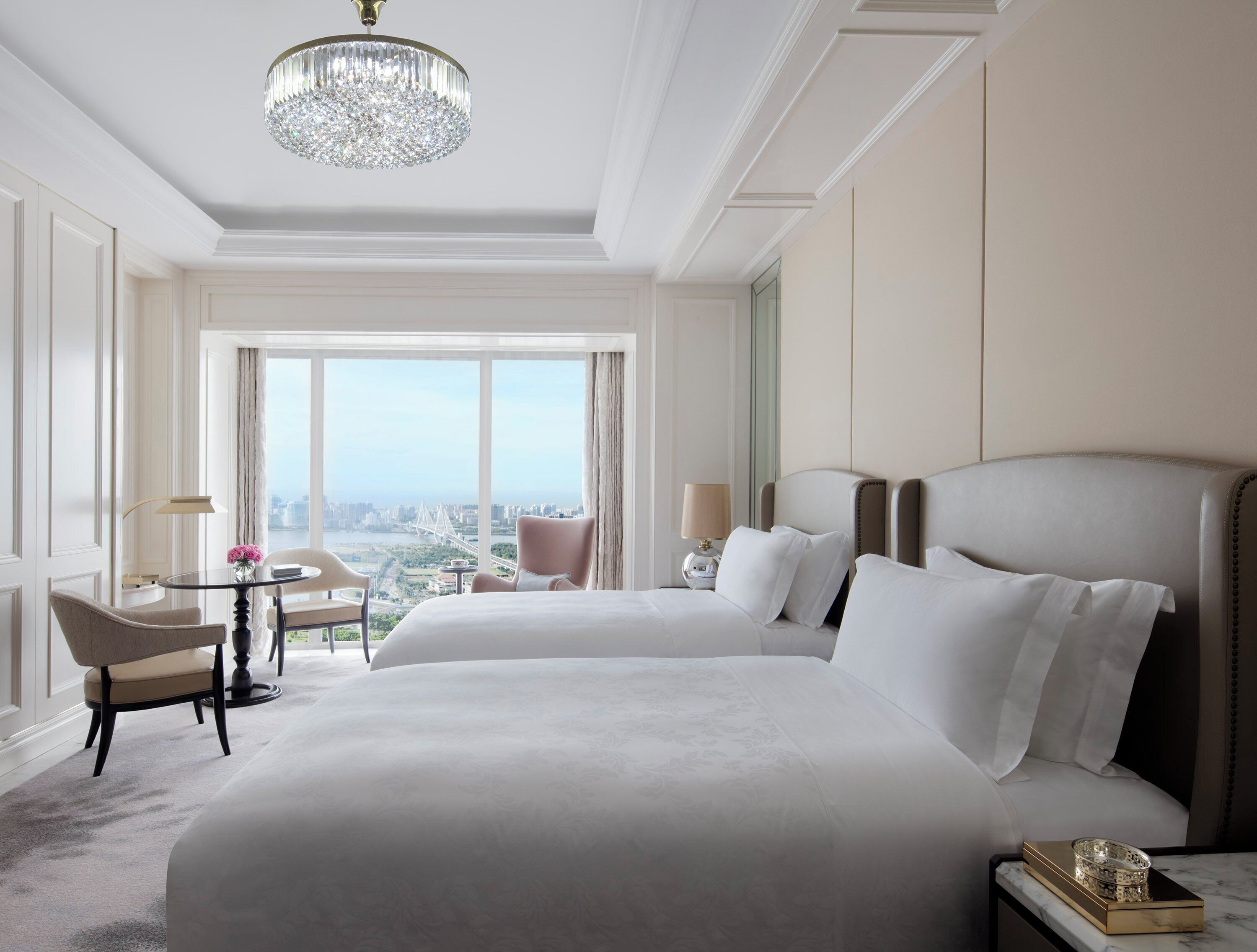 A hotel room with a large bed, a sitting area with a table and chairs, a chandelier, and a view of the city.