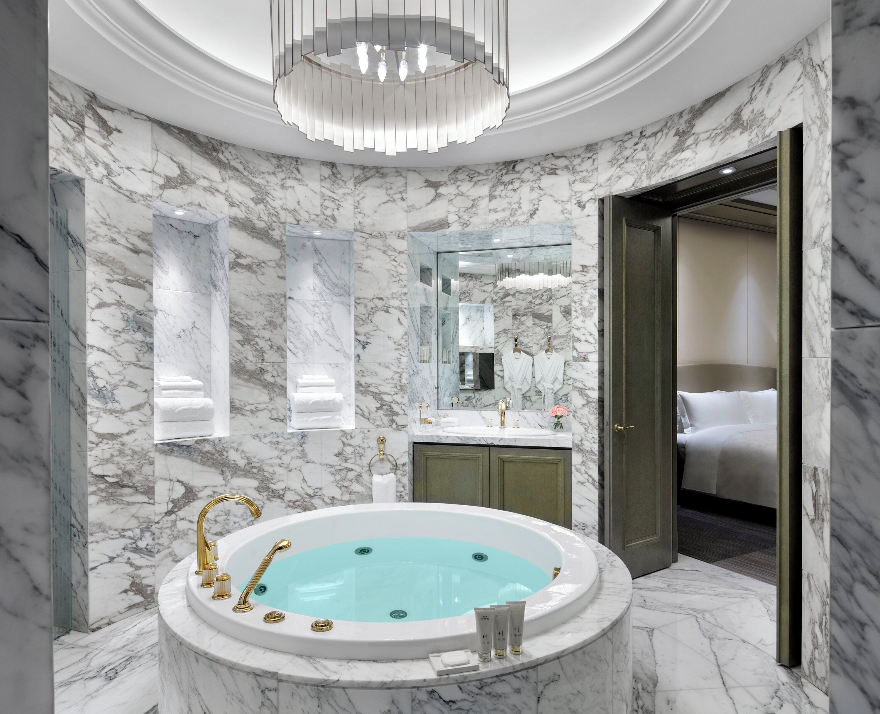 A luxurious bathroom with a marble floor, a freestanding bathtub, a large mirror, and a chandelier.