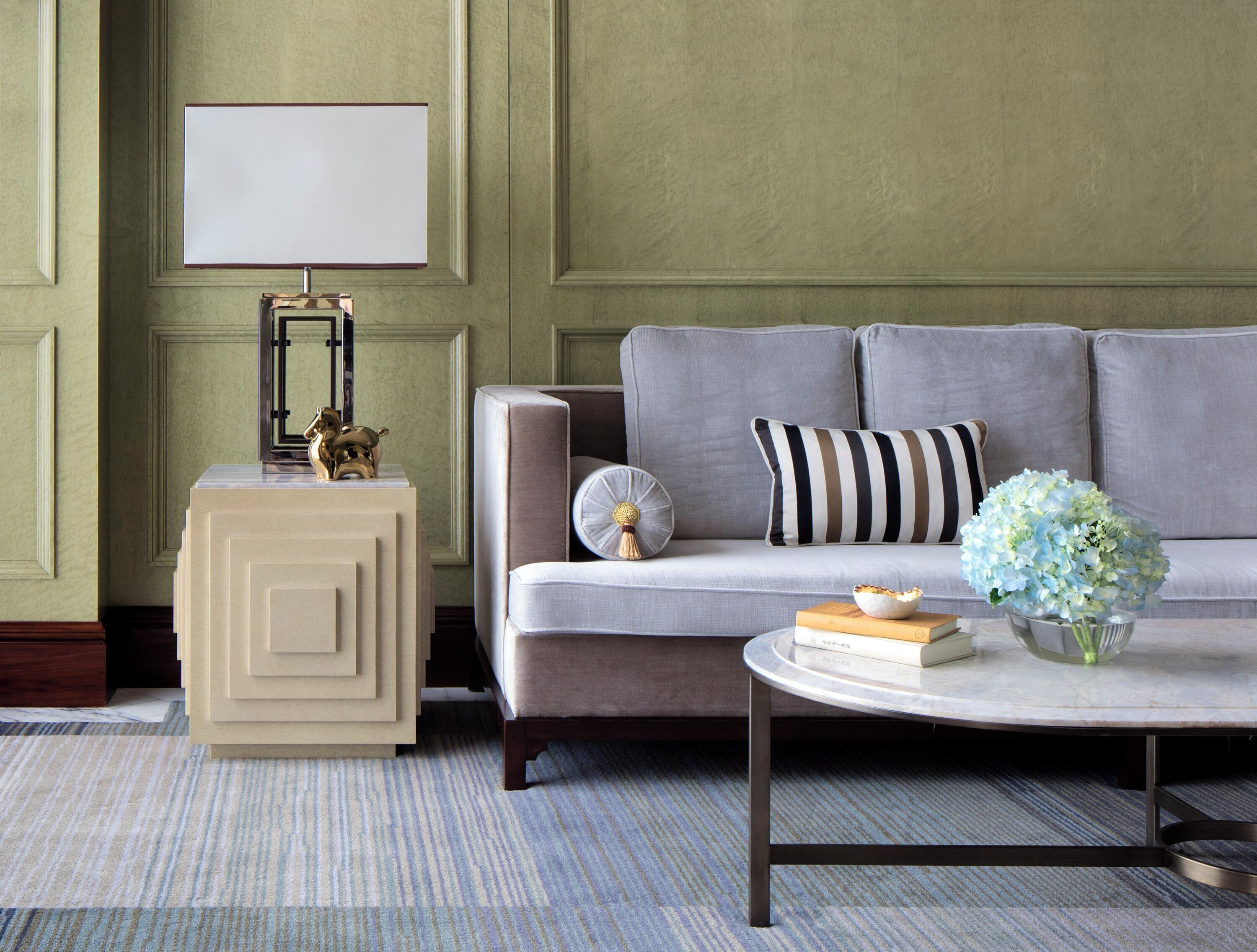 A living room with a grey sofa, a round coffee table with books and a vase, a side table with a lamp, and a framed picture on the wall.