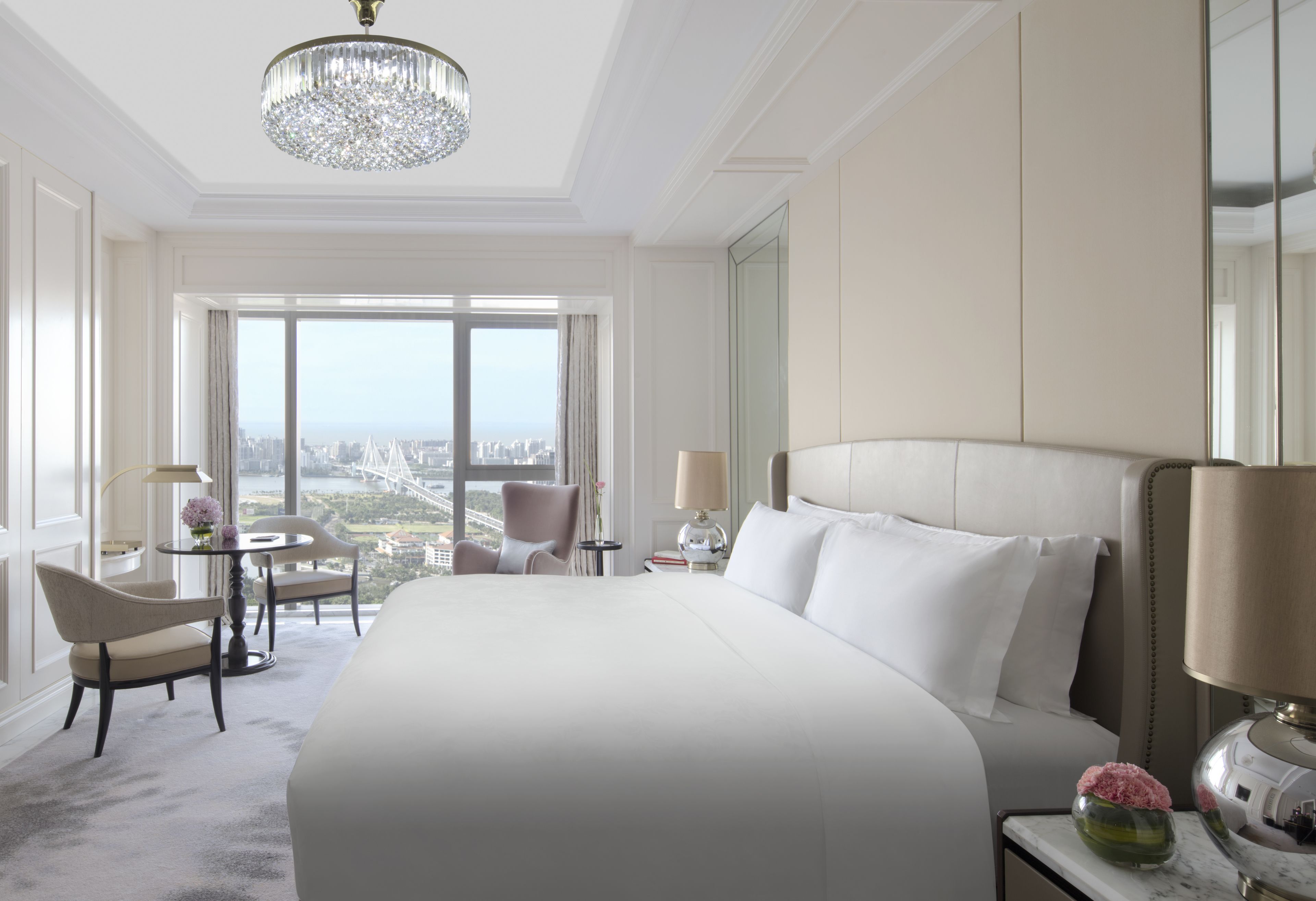 Camera d'albergo con un letto grande, un lampadario, vista sulla città, un tavolo rotondo con sedie e un tavolino con una lampada.