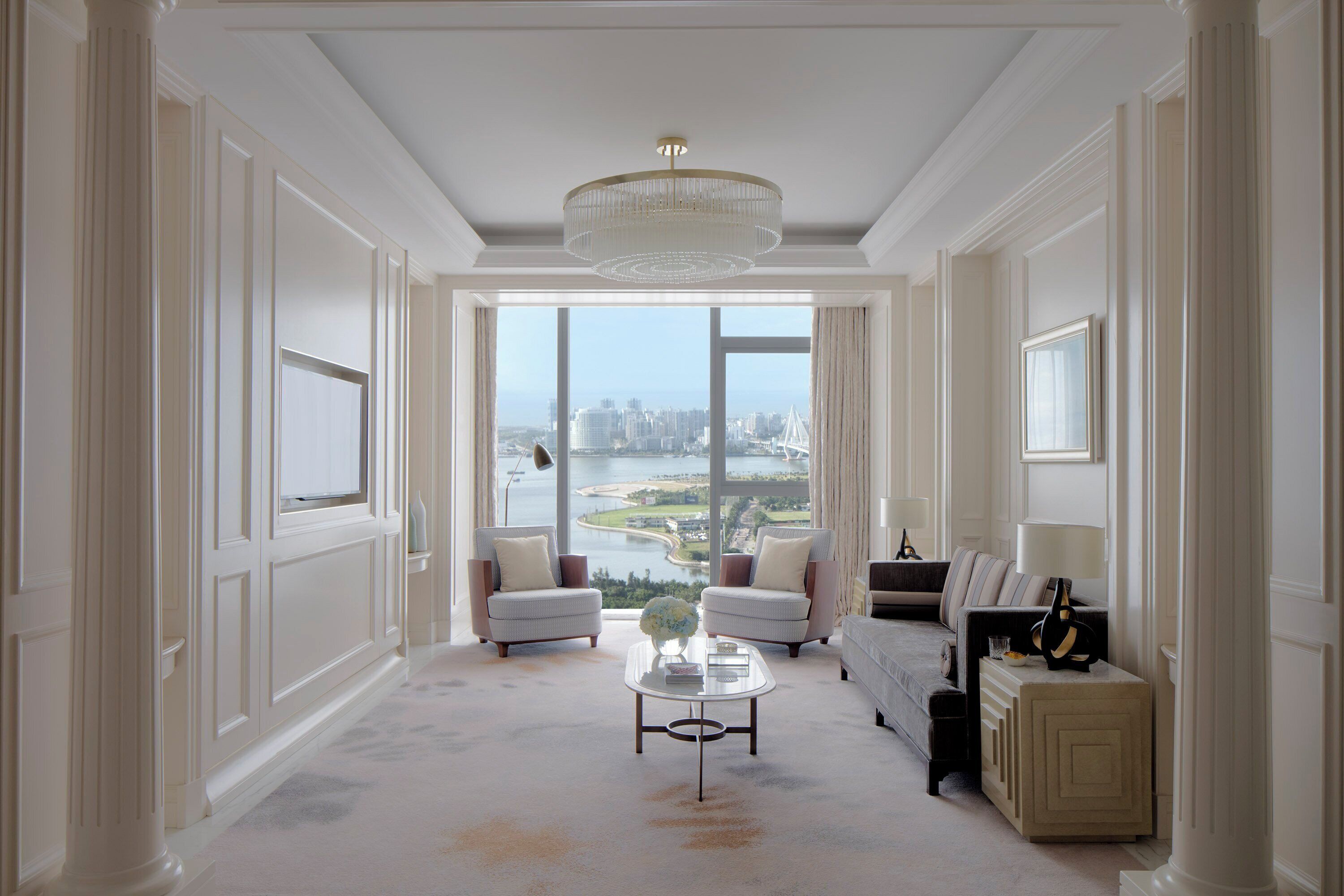 Un salon spacieux doté d’une grande fenêtre offrant une vue sur la ville, d’un lustre et d’une table basse moderne.