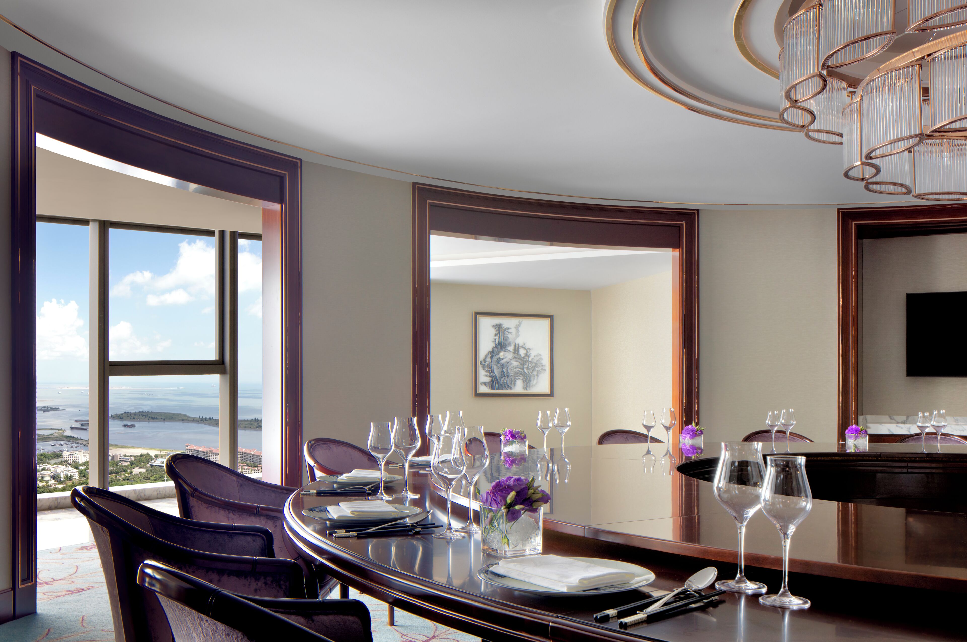 Un comedor con vistas al mar, una mesa dispuesta con copas de vino y platos, y una araña.
