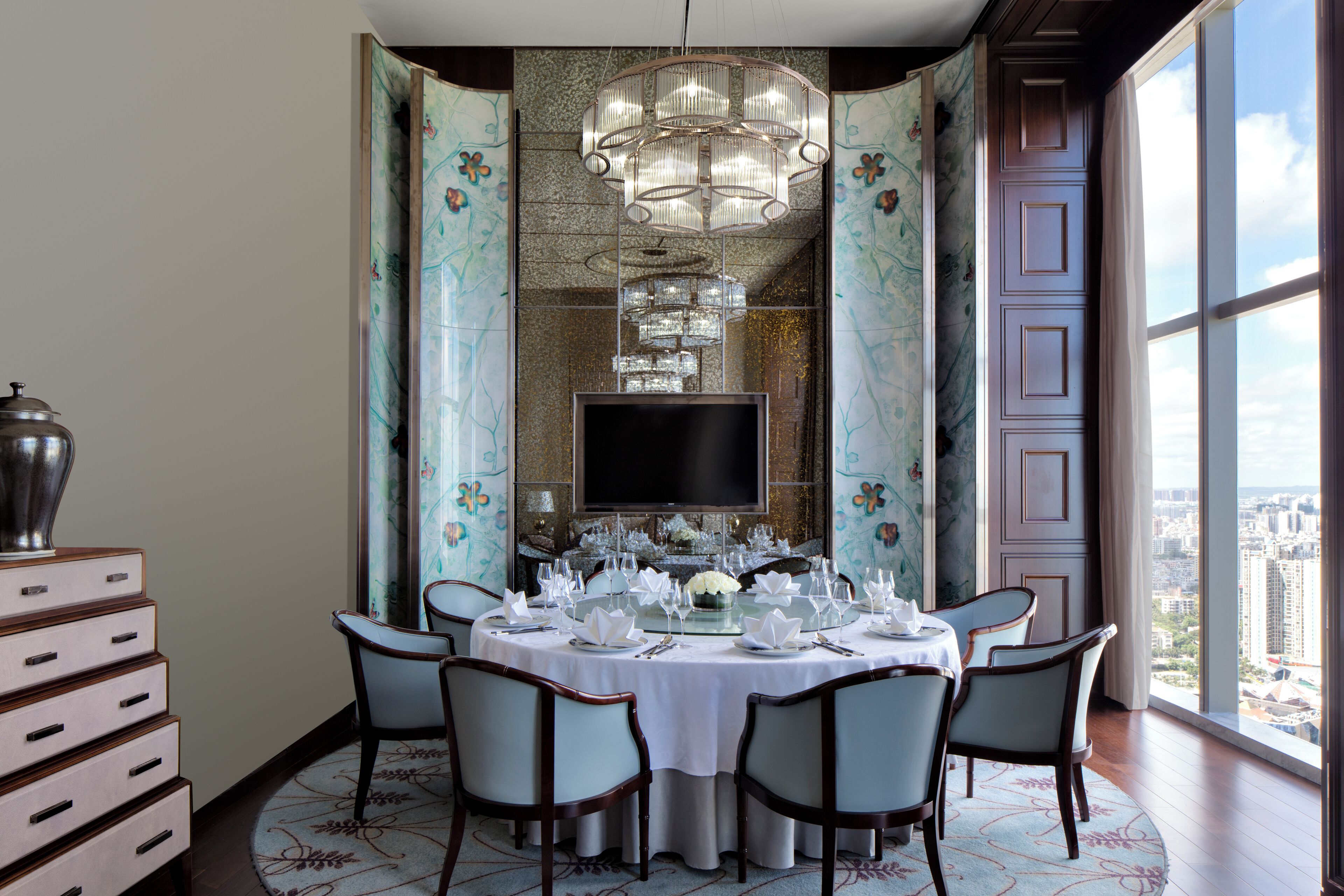 A dining area with a round table set for six, surrounded by chairs, a chandelier, and a large window offering a city view.
