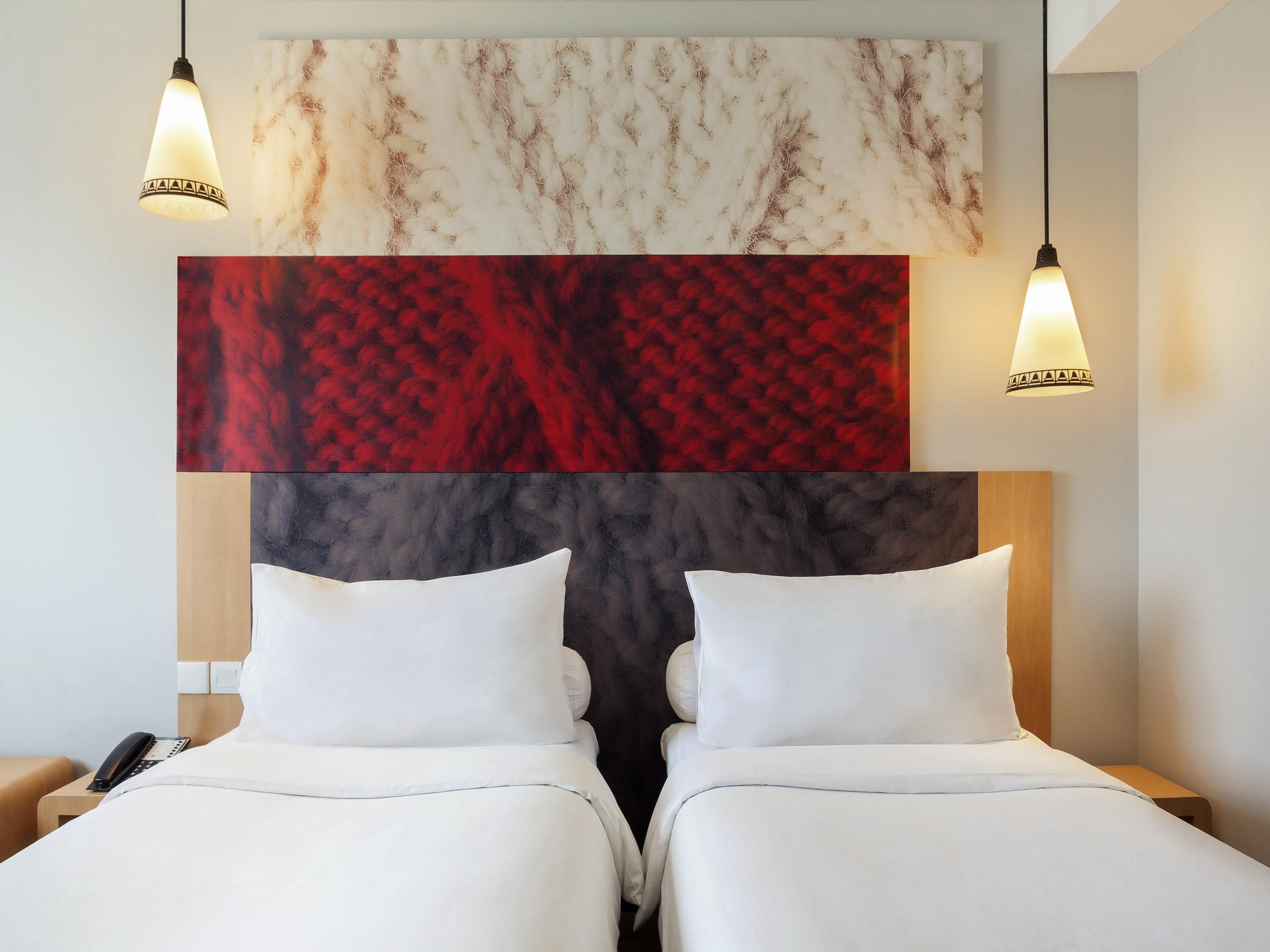 A hotel room with two beds, a large abstract painting above the headboard, and two pendant lights.