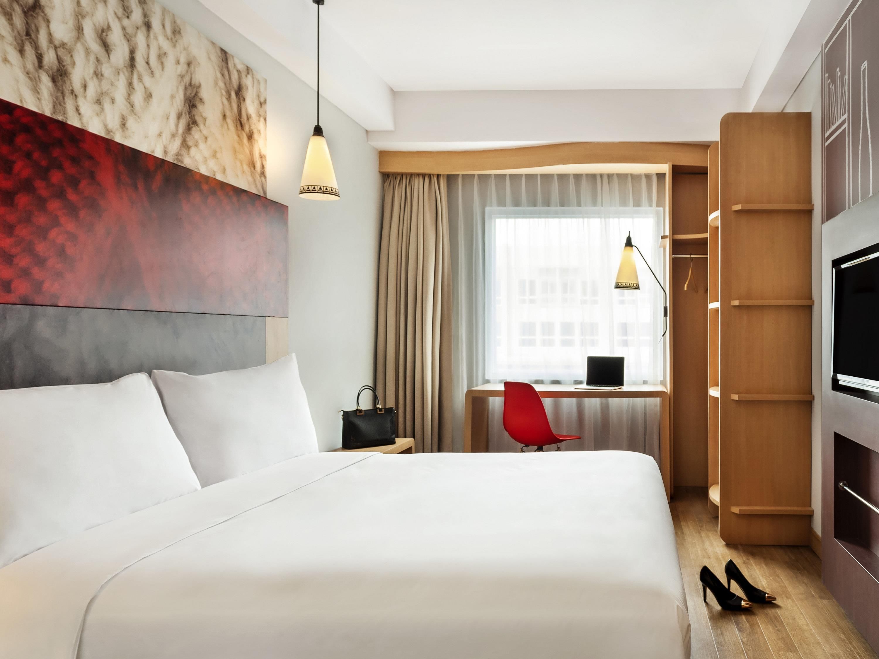 A hotel room with a large bed, a desk with a laptop, a red chair, a TV, and a wardrobe.