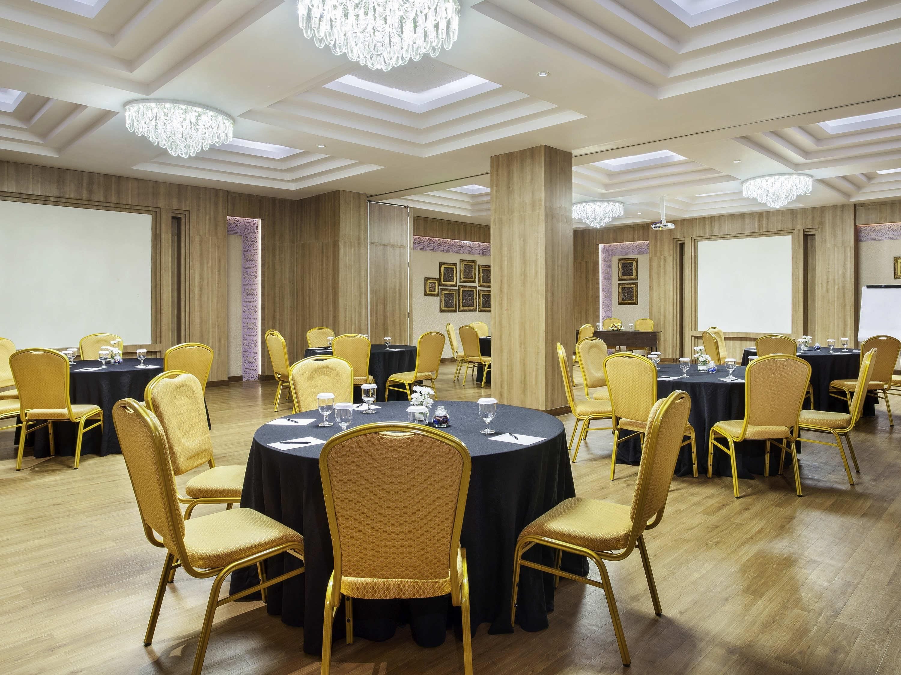 Une salle de conférence dotée de tables rondes, de chaises dorées, de lustres et d’un plancher en bois.