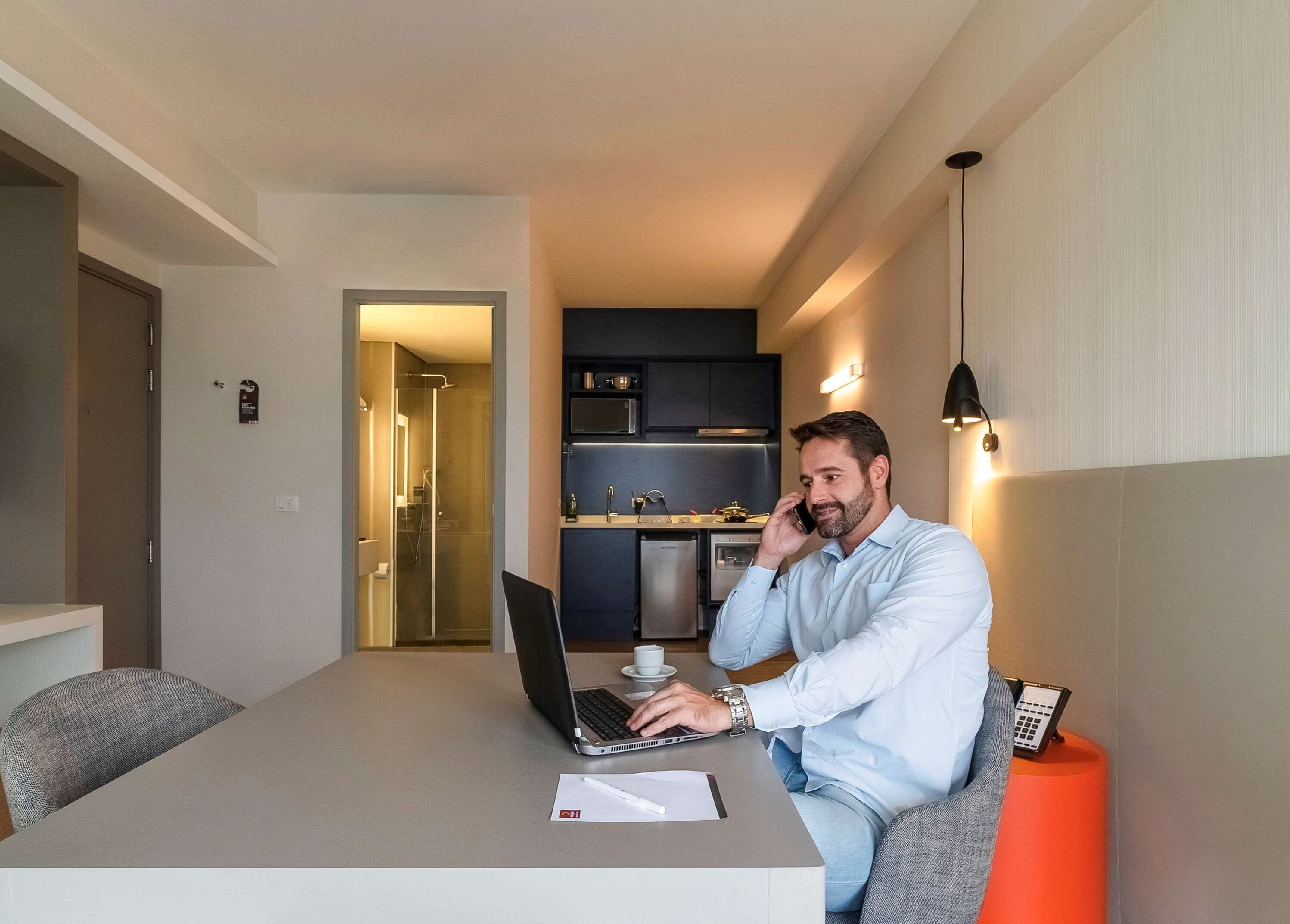 Un hombre sentado a una mesa con una computadora portátil, hablando por teléfono. Se observa una cocina al fondo con electrodomésticos modernos.