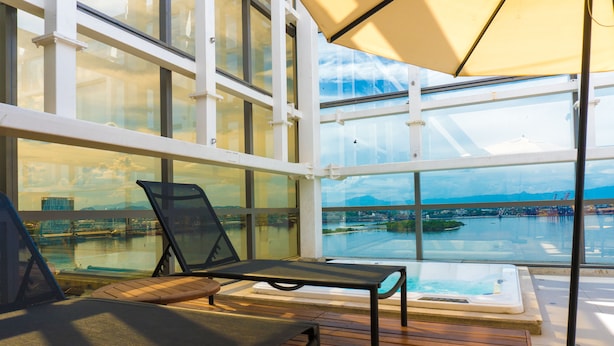 A sunlit deck with two lounge chairs, a hot tub, and a glass-enclosed view of a body of water and distant landscape. - Novotel RJ Porto Atlântico (Rio de Janeiro)