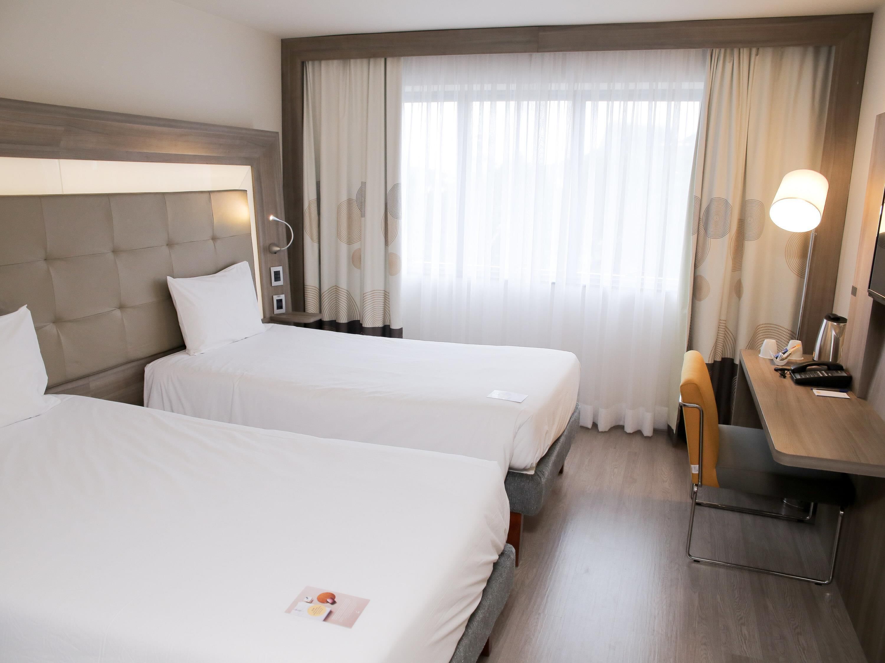 A hotel room with two beds, a desk, a chair, a lamp, and a window with curtains.
