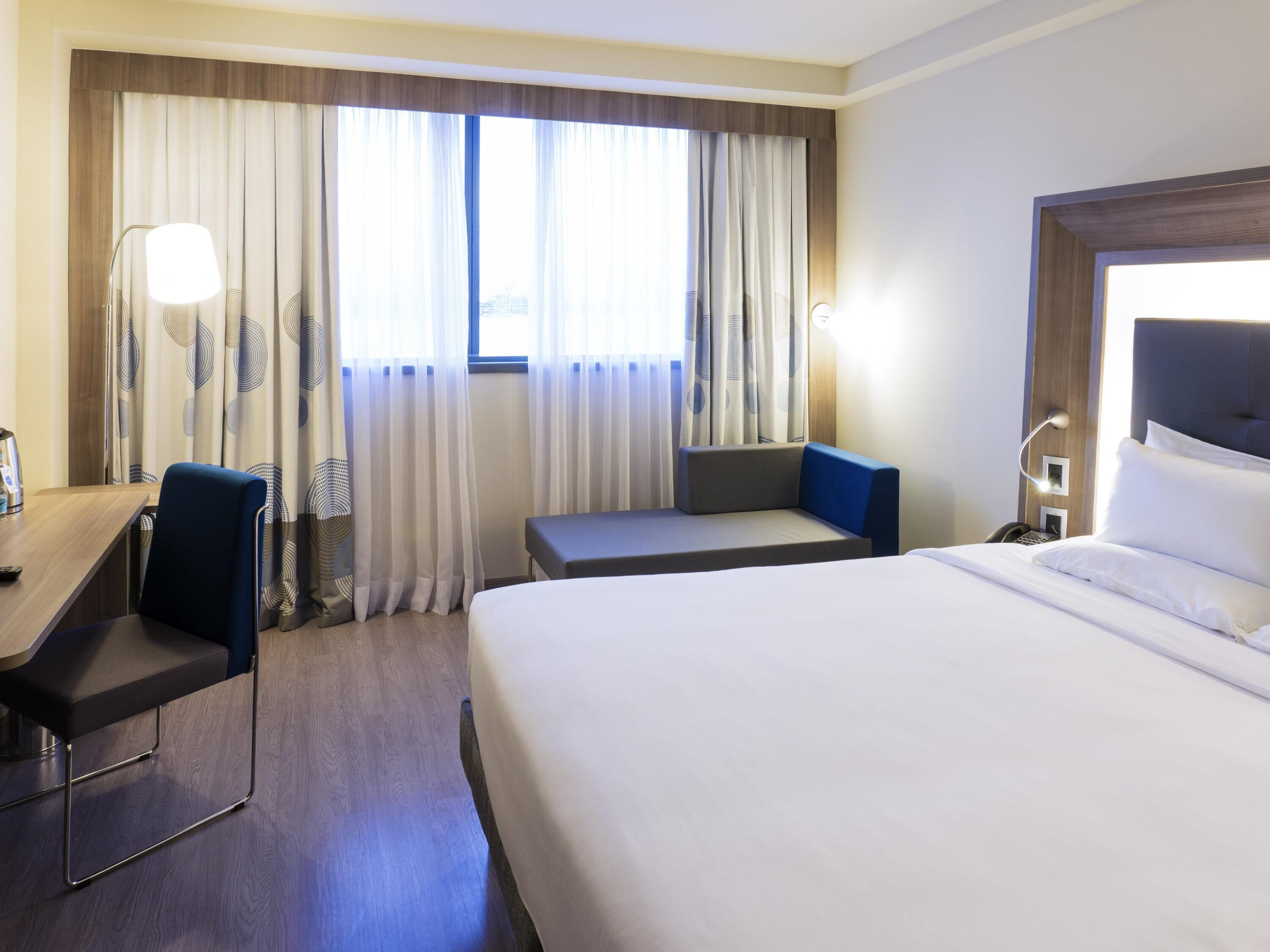A hotel room with a bed, a desk, a chair, a small table, a lamp, and curtains.