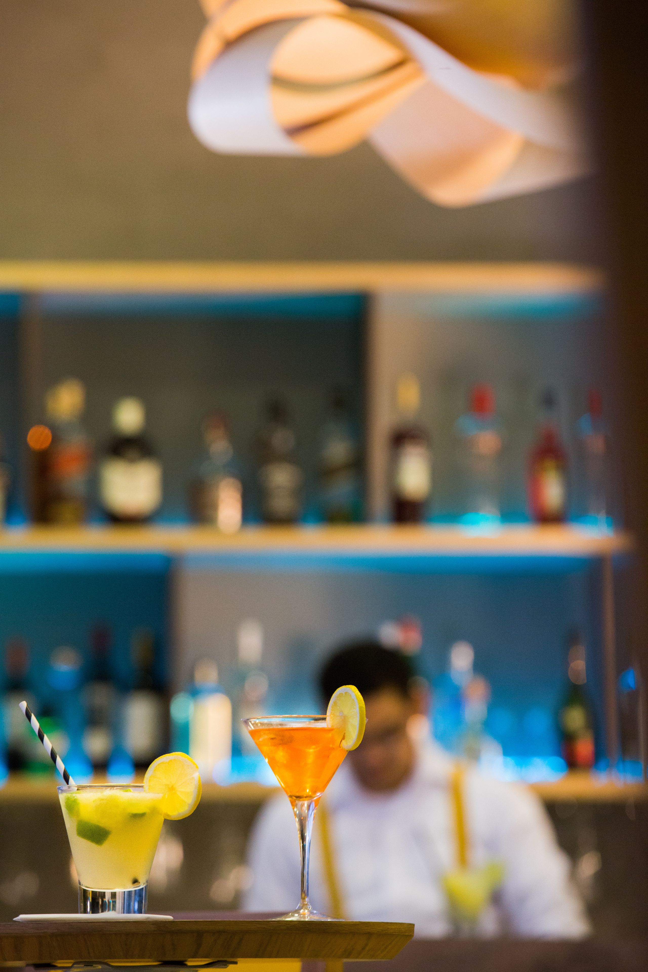 A bar setting with two cocktails on a tray, a bartender in the background, and shelves with bottles.