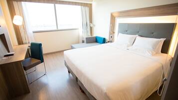 Une chambre d’hôtel avec un grand lit, un bureau avec une chaise, une petite table et une fenêtre avec des rideaux.