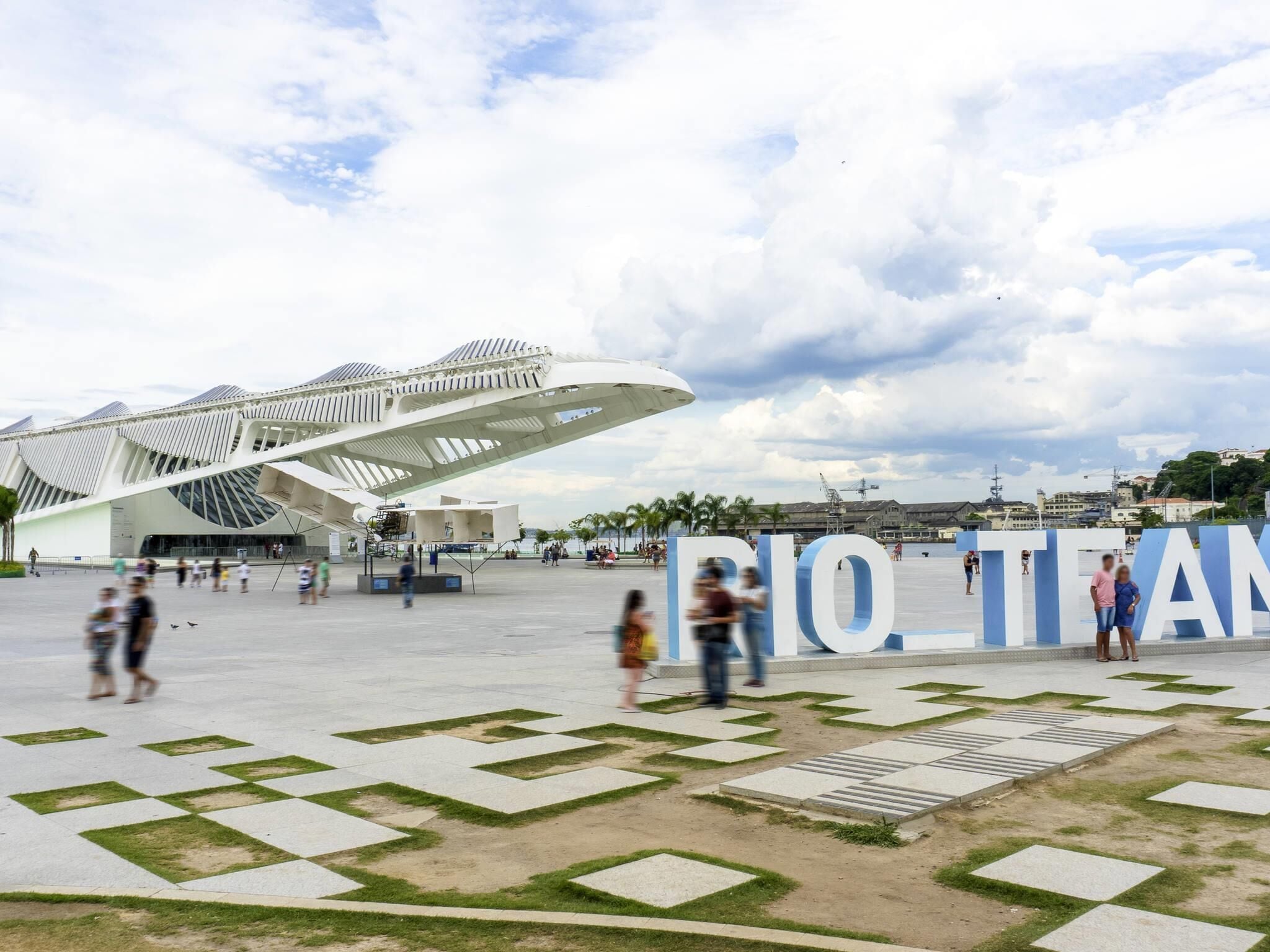 A modern architectural structure with a unique design, surrounded by a spacious plaza with geometric patterns and a large sign reading "RIO TEAM".