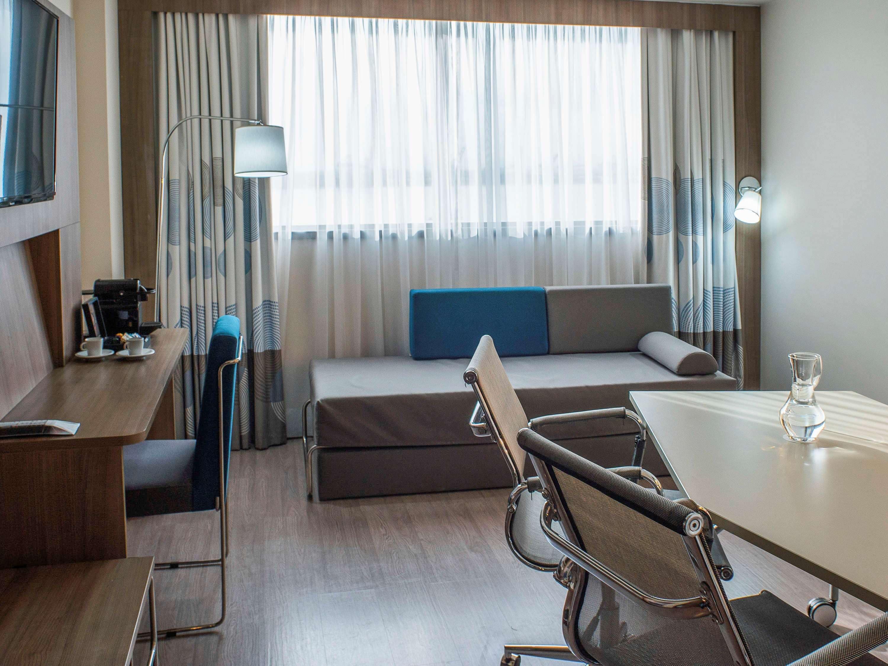 A hotel room with a bed, a desk, a chair, a coffee machine, and a lamp.