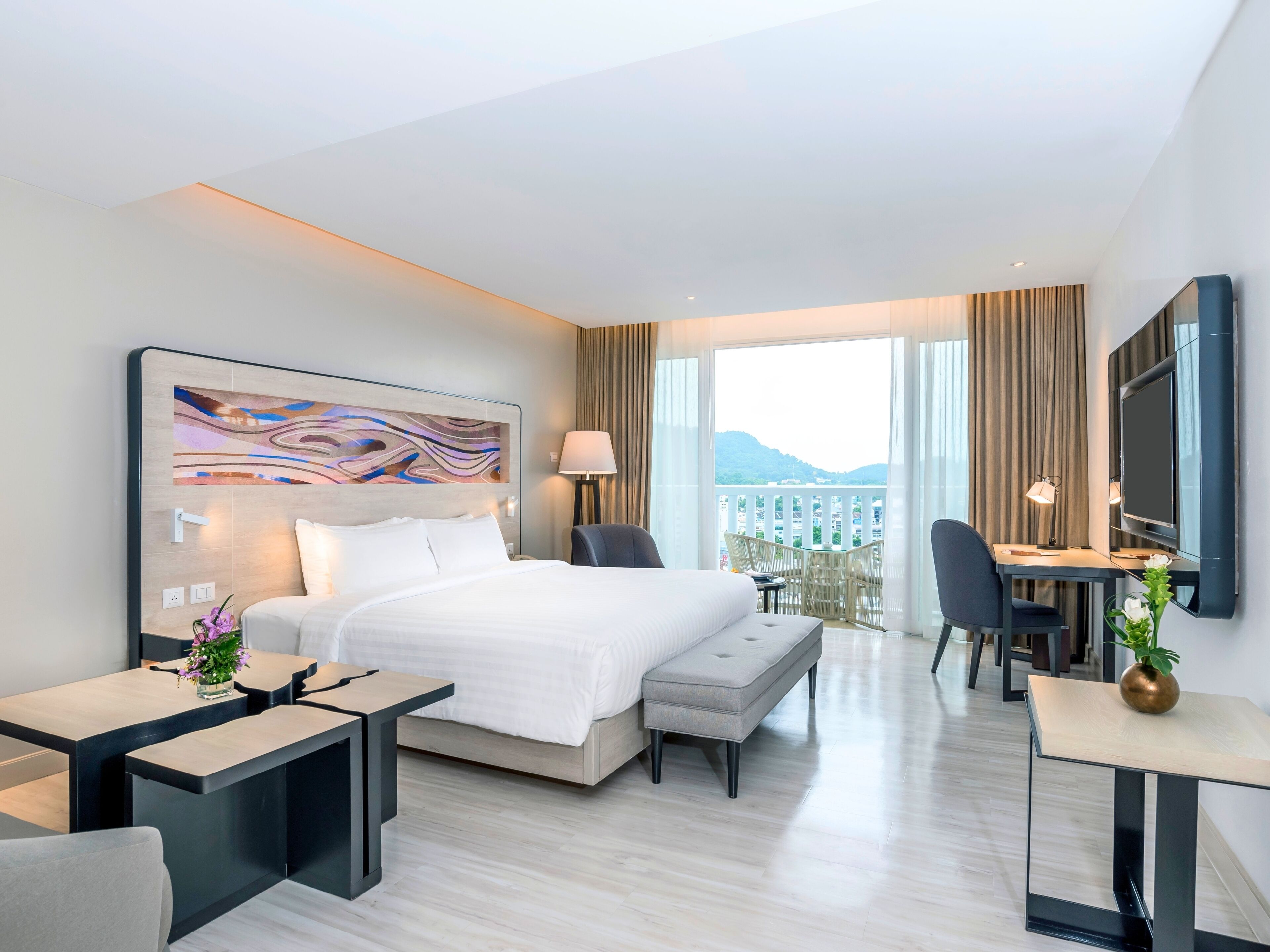 A modern hotel room with a large bed, a desk, a chair, and a balcony with a view of the mountains.