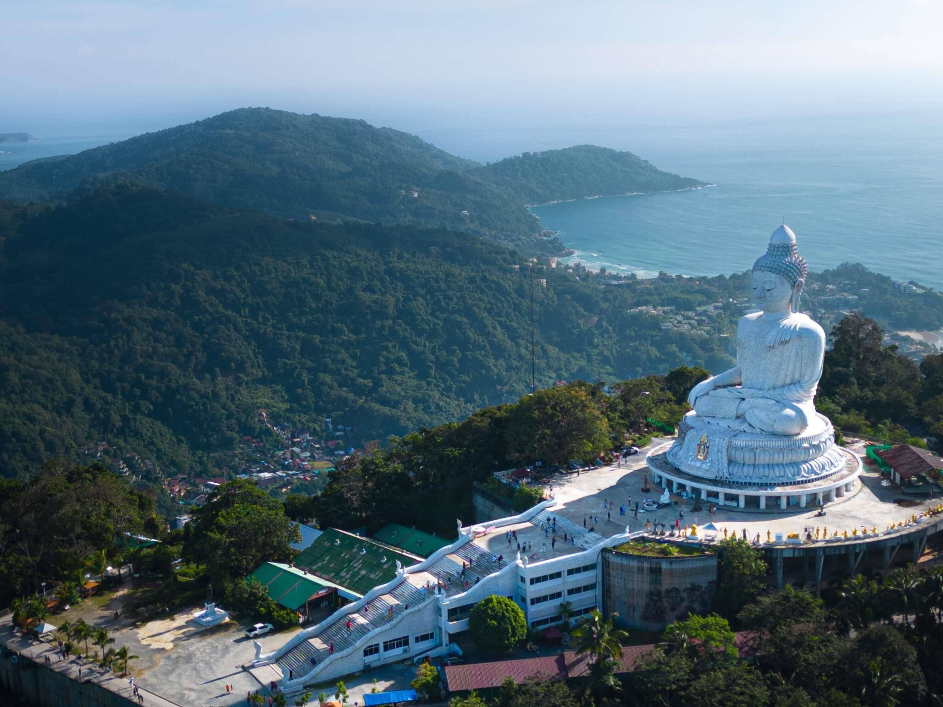 A large white Buddha statue surrounded by a circular walkway, with a mountainous backdrop and a view of the sea.